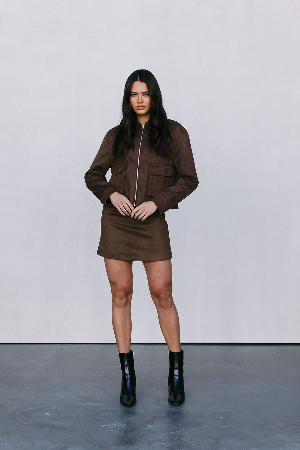 Woman wearing a brown outfit with a jacket and skirt, standing against a plain background.