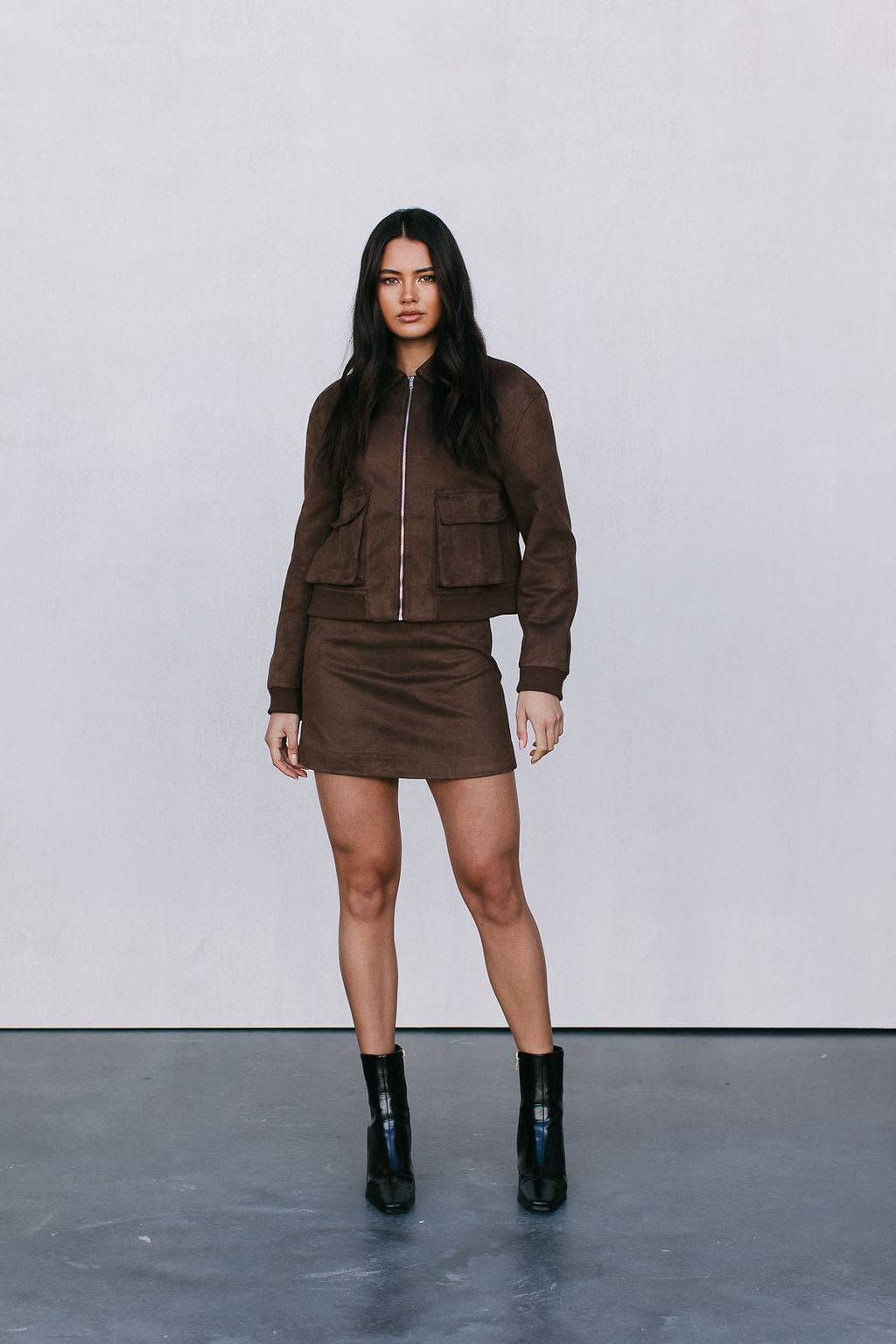 Woman wearing a brown jacket and skirt set with black boots against a plain background