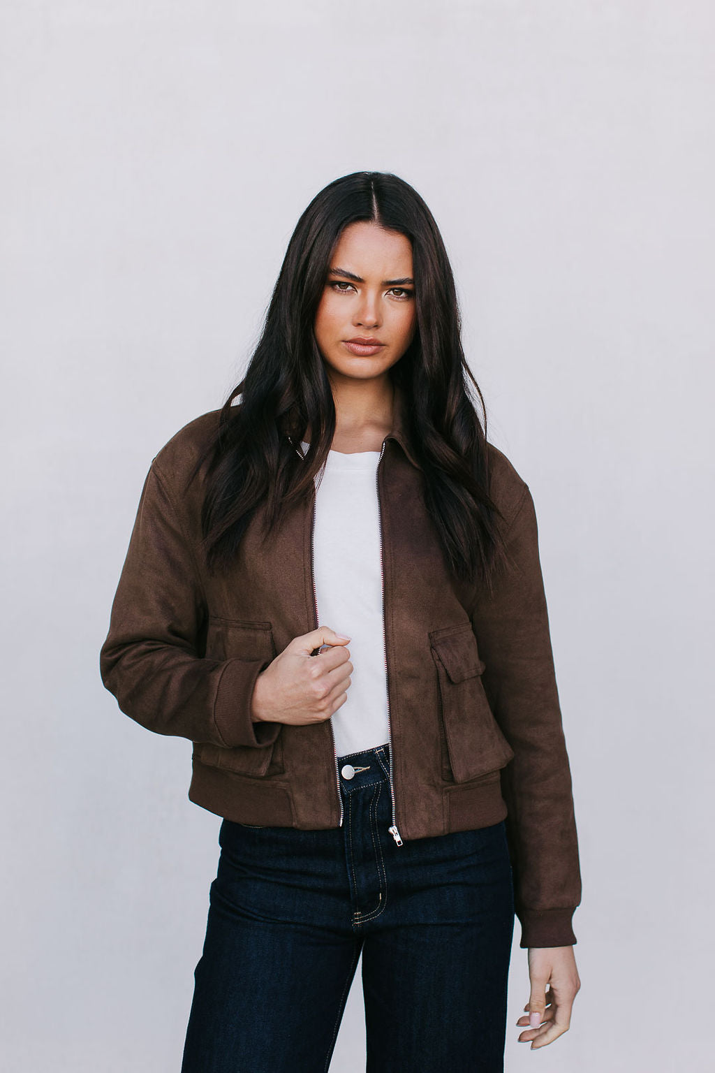 Woman wearing a brown suede bomber jacket over a white shirt and dark jeans on a plain background