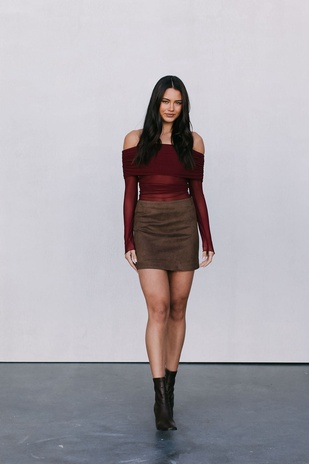 Woman wearing a burgundy off-shoulder top and brown suede skirt against a white background