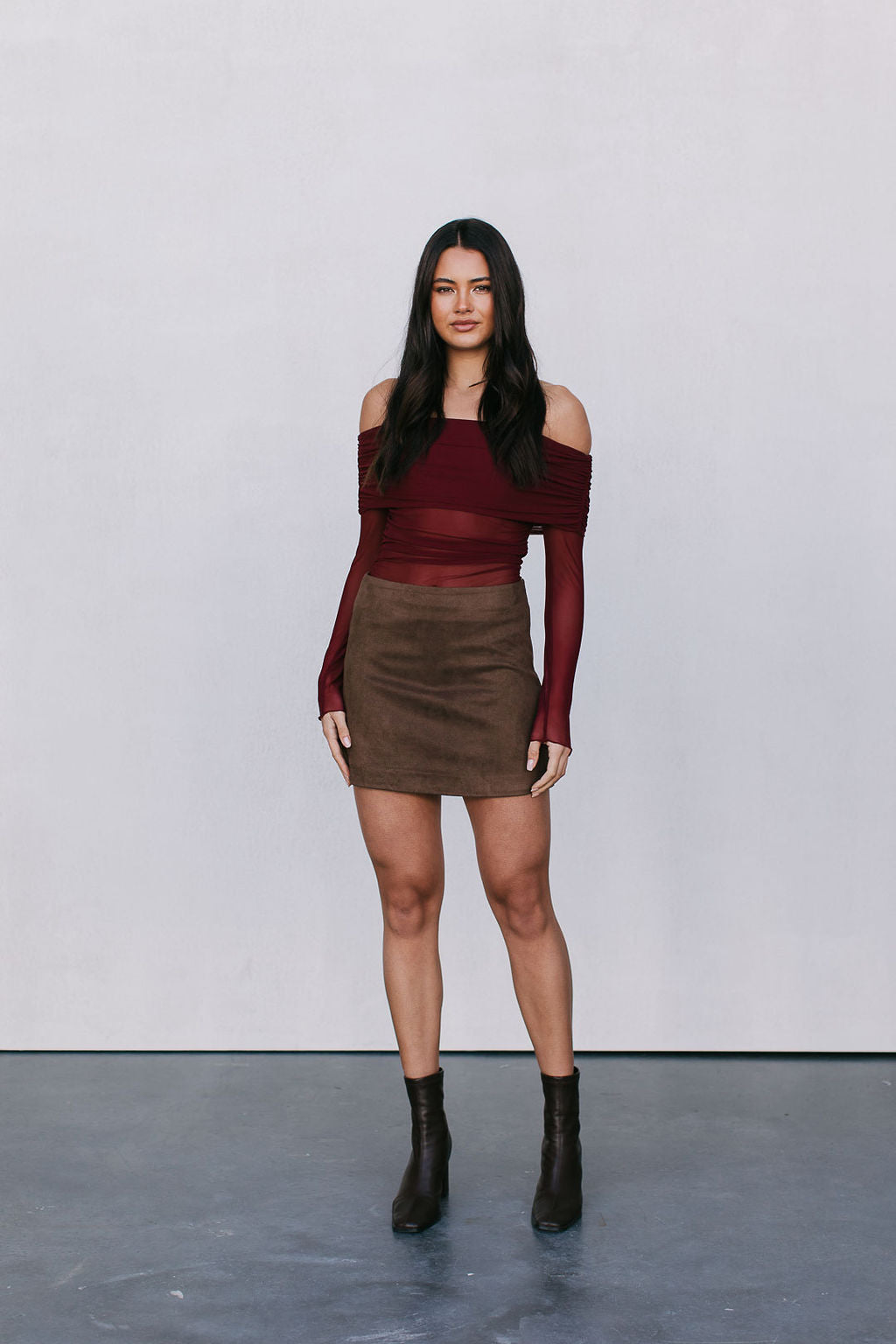 Woman wearing a burgundy off-shoulder top and brown skirt against a plain background