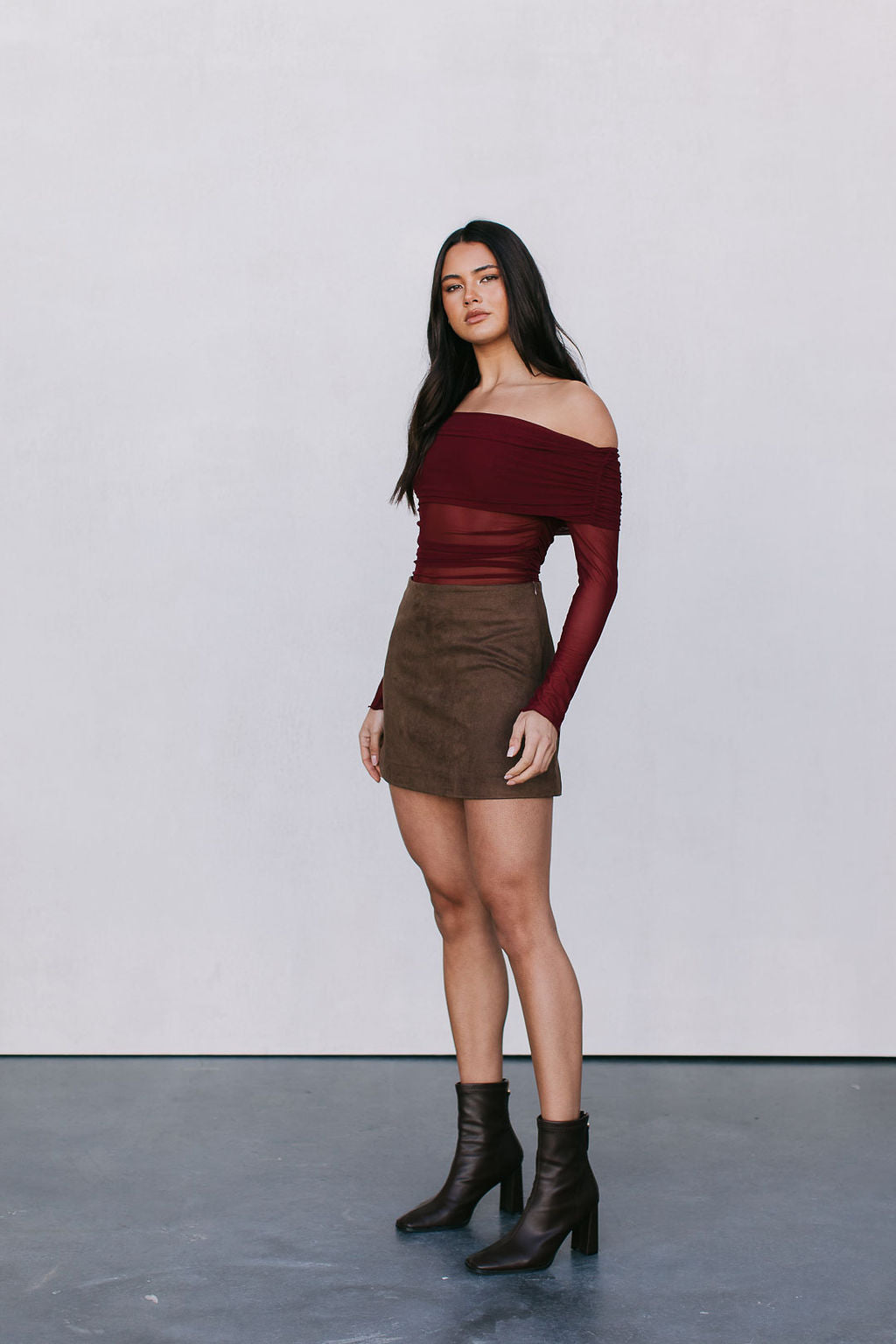 Woman wearing a burgundy off-shoulder top and brown skirt with black boots against a white background