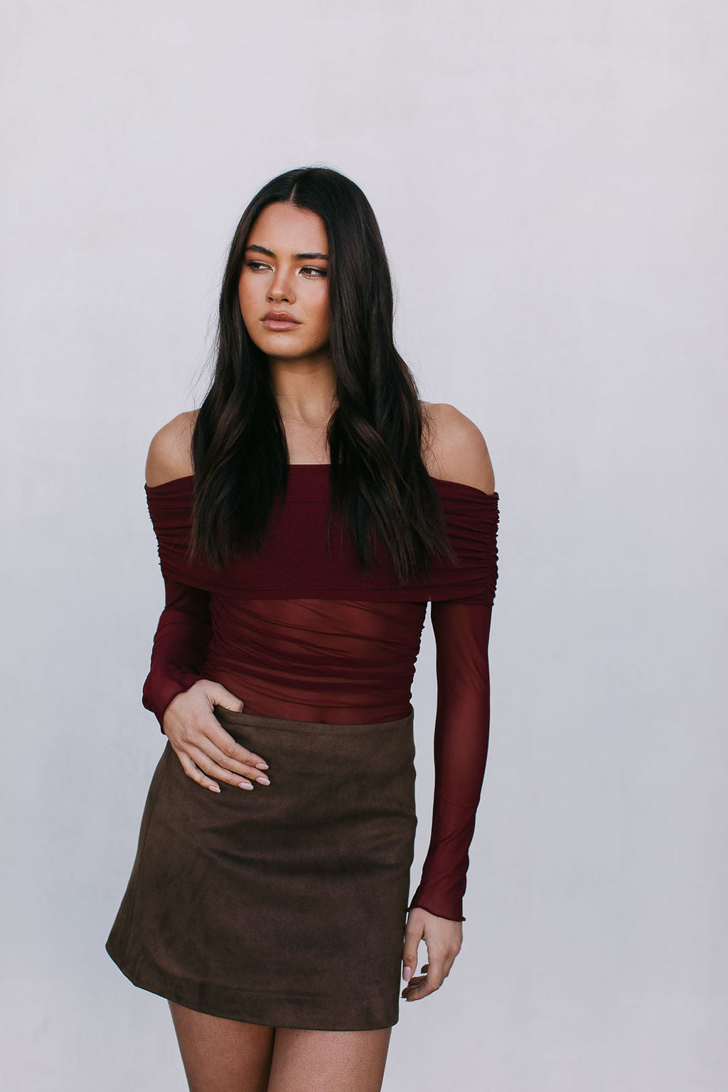 Woman wearing a burgundy off-shoulder top and brown suede skirt against a plain background