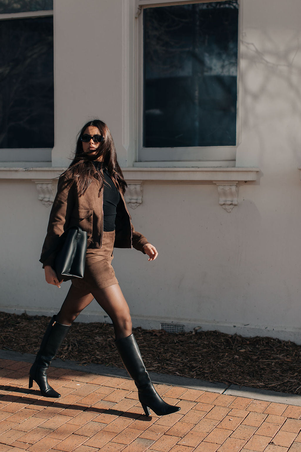 Woman walking on a brick sidewalk wearing a brown suede bomber jacket, black top, tan pants, and black knee-high boots.