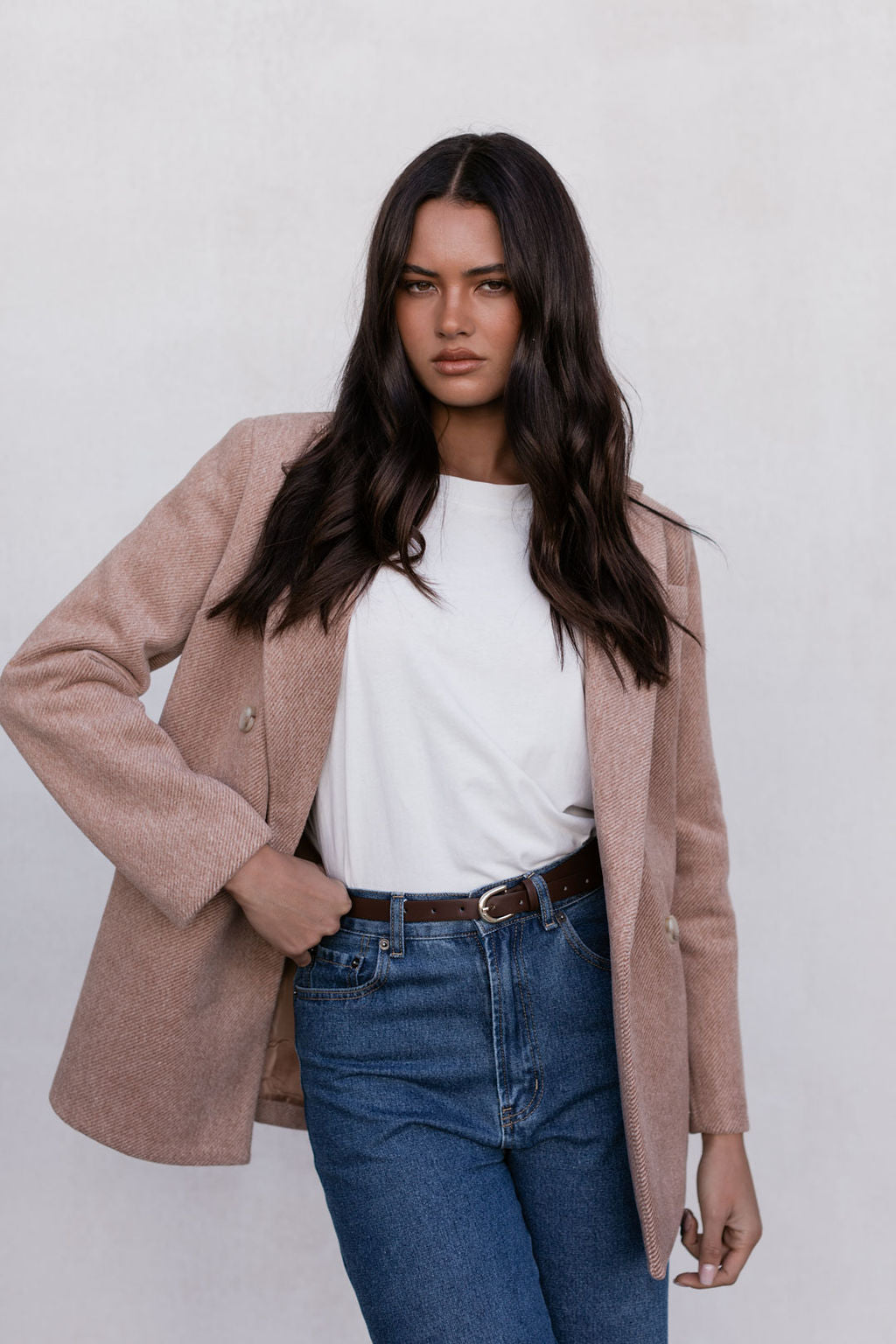 Woman wearing a beige coat, white shirt, and blue jeans against a plain background