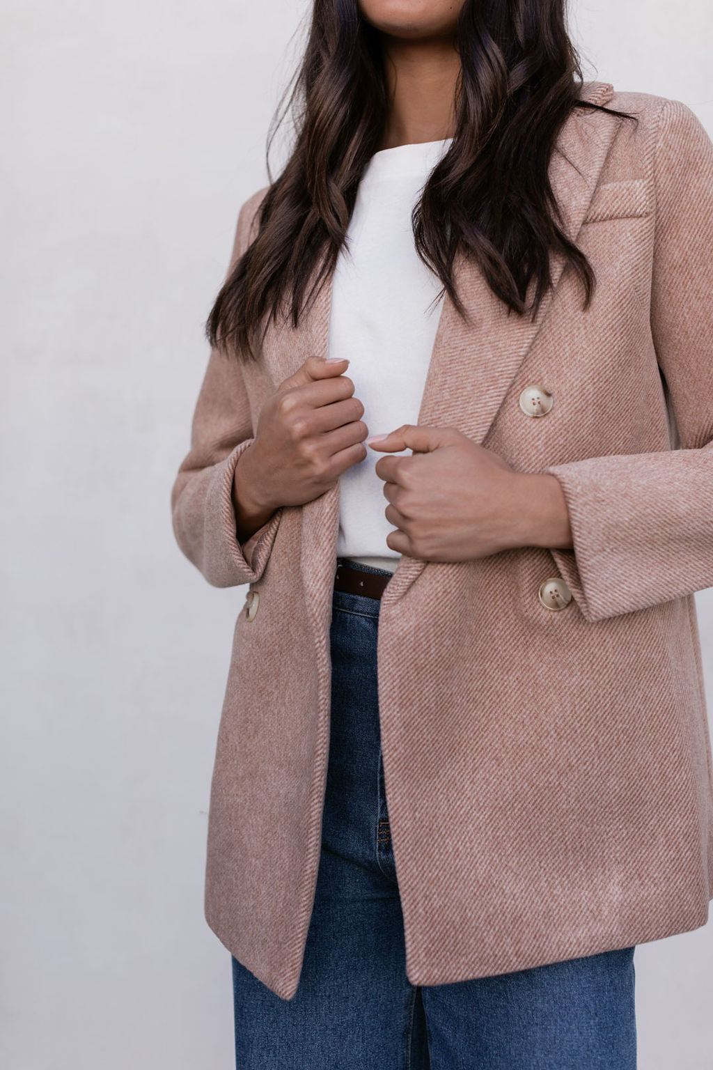 Person wearing a beige blazer over a white top and blue jeans on a light gray background