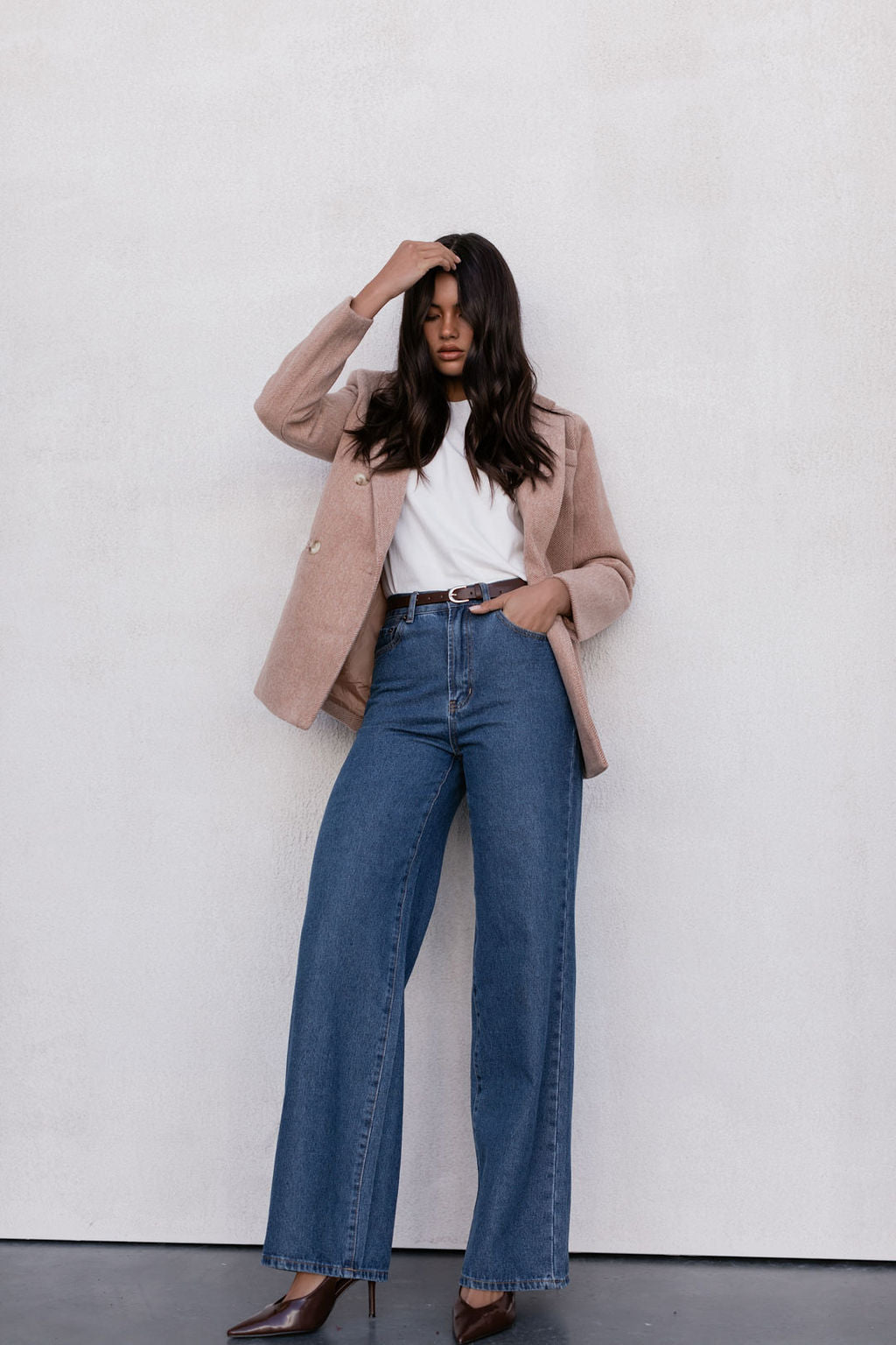 Woman wearing a beige blazer, white shirt, and blue jeans against a white wall.