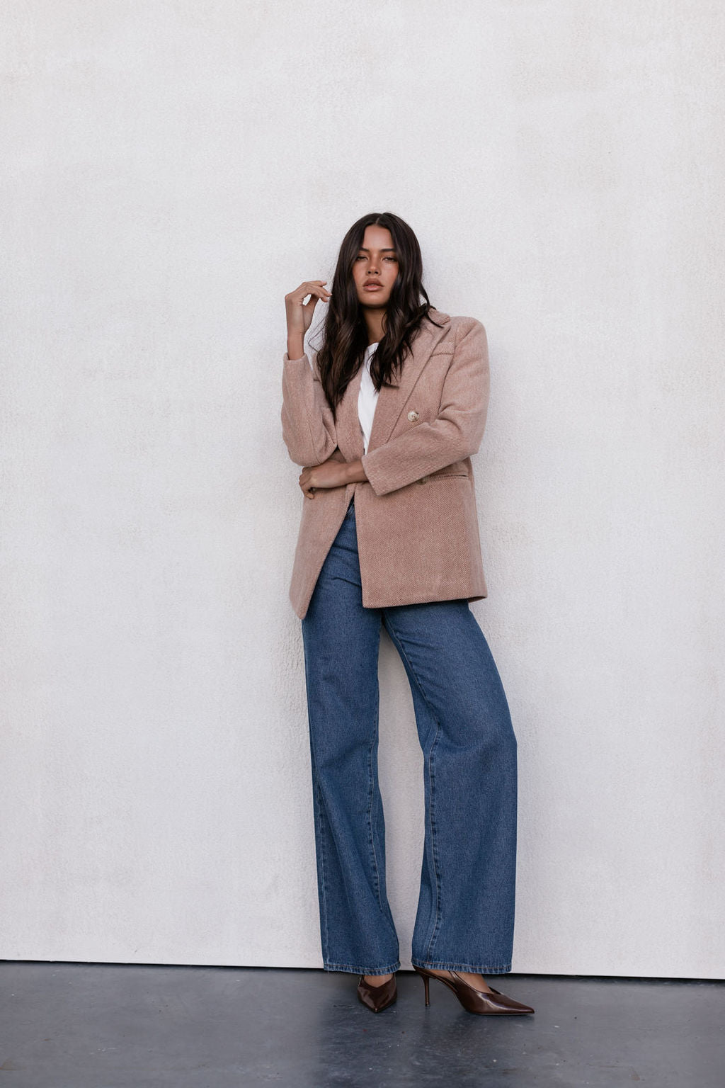 Woman wearing a beige blazer, white top, and blue jeans against a white wall.