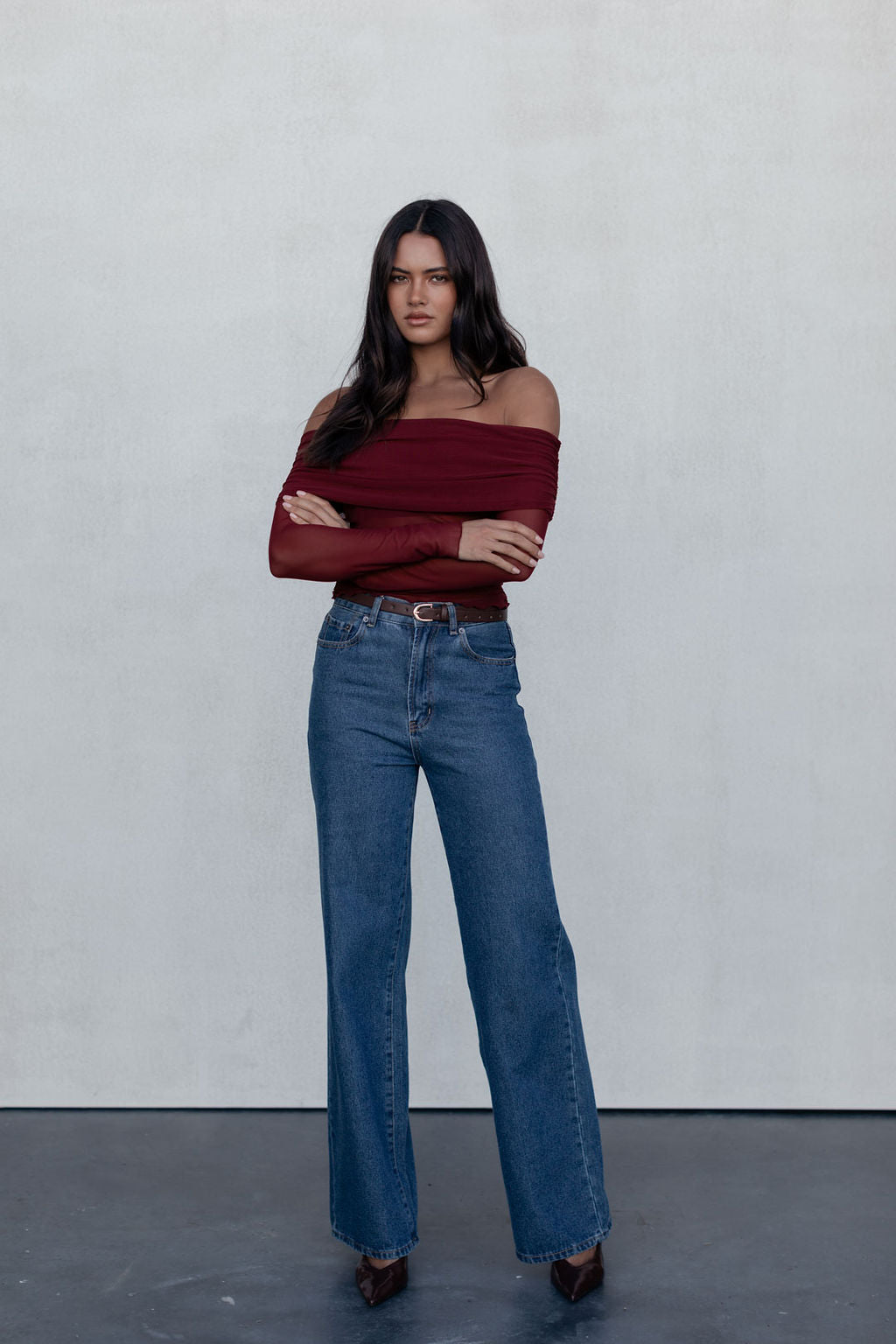 Woman wearing a burgundy off-shoulder top and blue jeans against a plain background