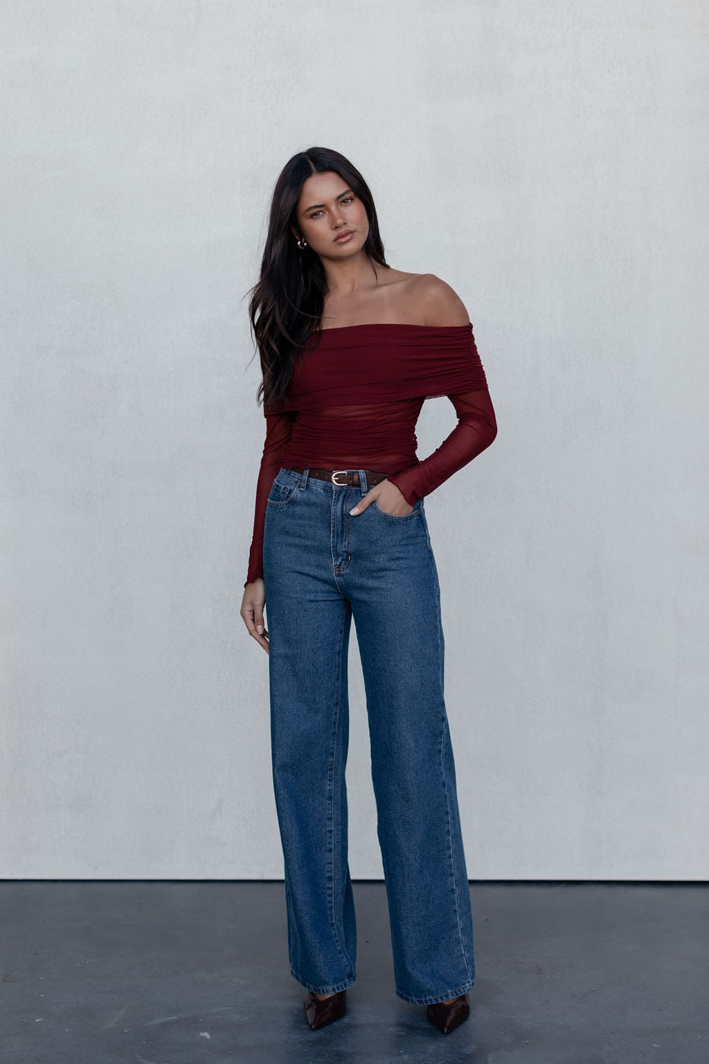 Woman wearing a burgundy off-shoulder top and blue jeans against a plain background