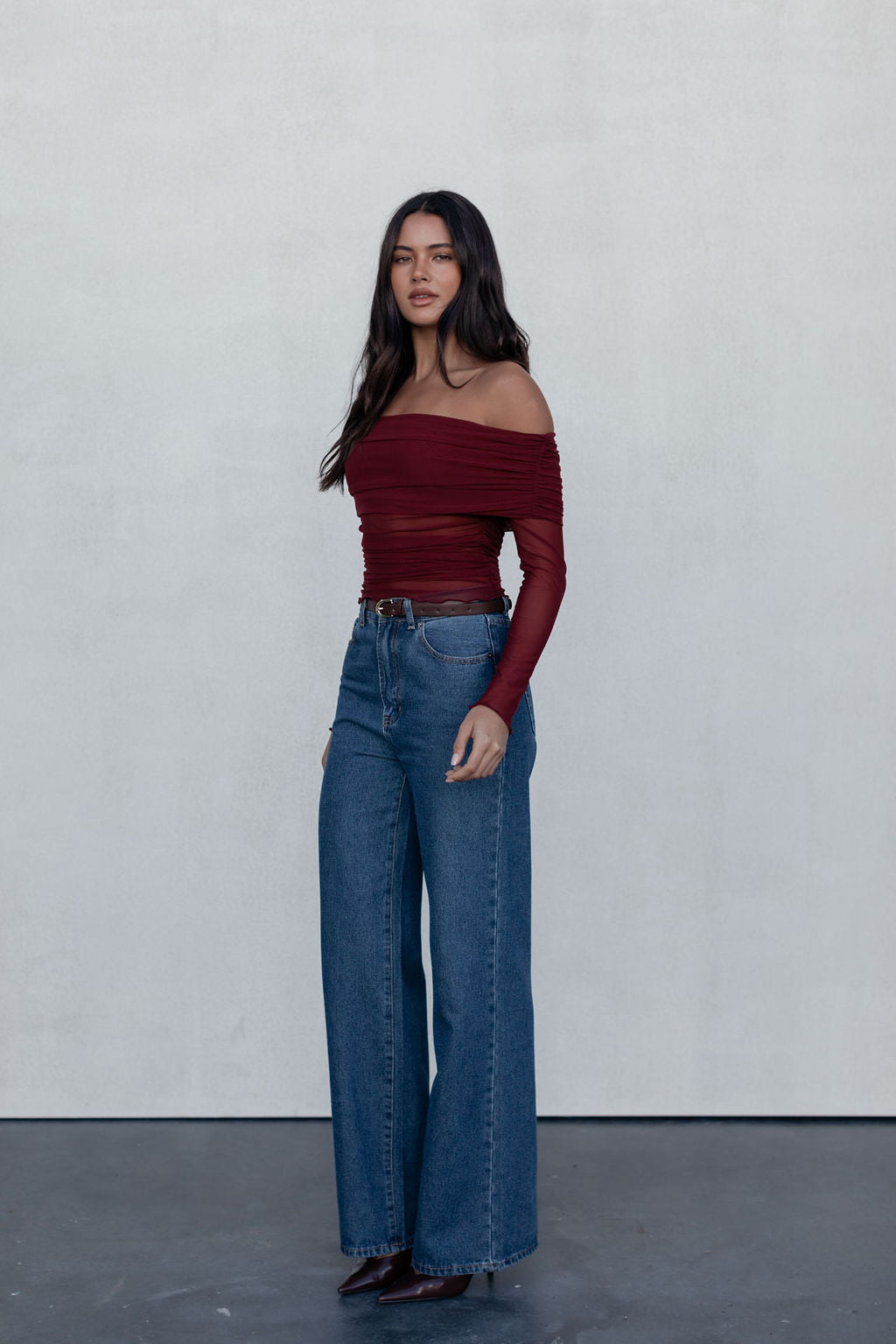Woman wearing a burgundy off-shoulder top and blue jeans against a white background
