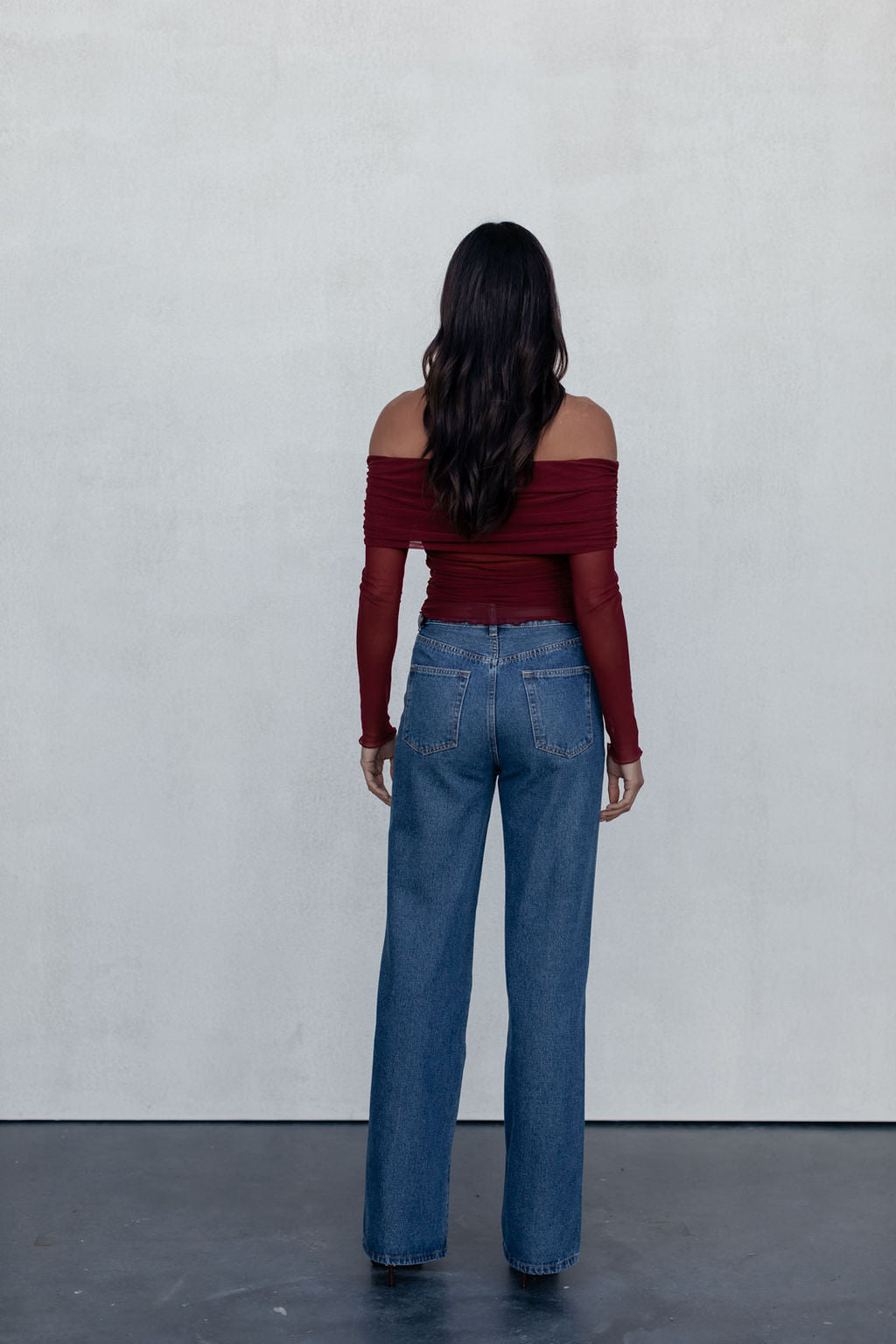 Woman wearing a red off-shoulder top and blue jeans against a white wall.