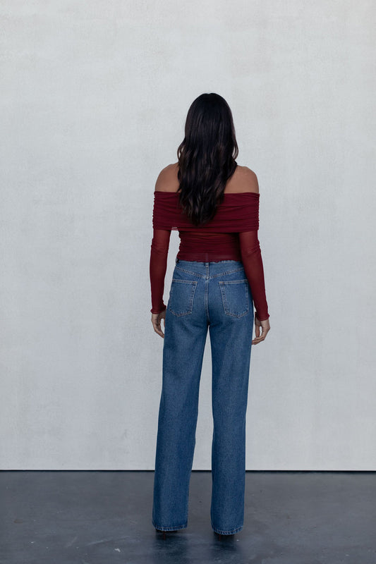 Woman wearing a red off-shoulder top and blue jeans against a white wall.