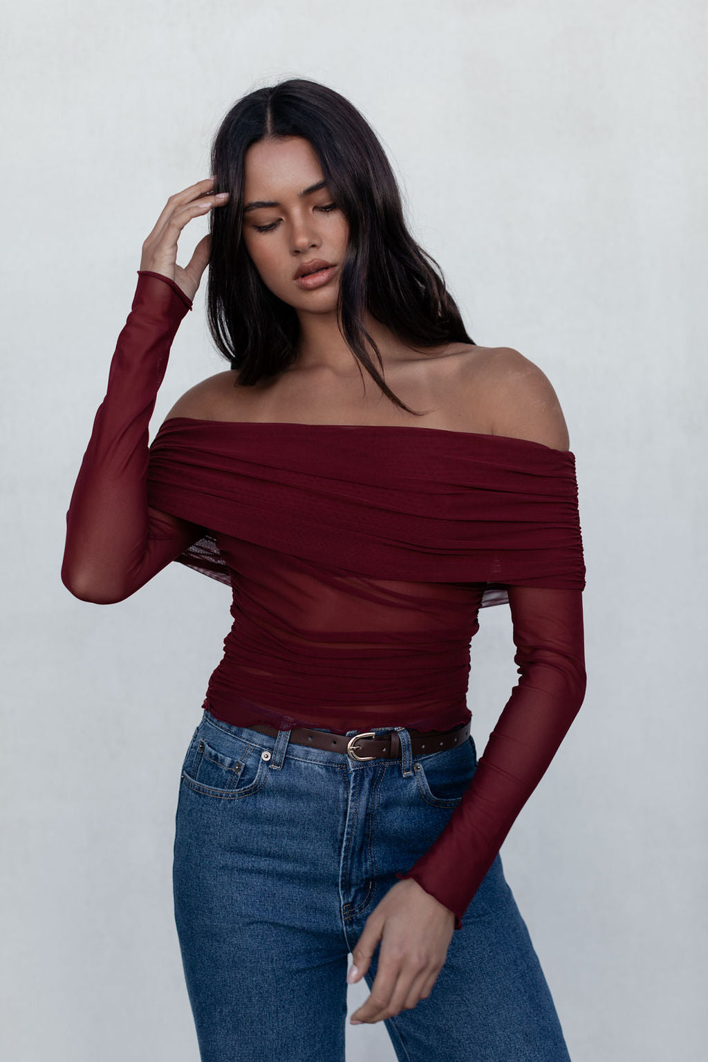Woman wearing a burgundy off-shoulder top and blue jeans against a white background