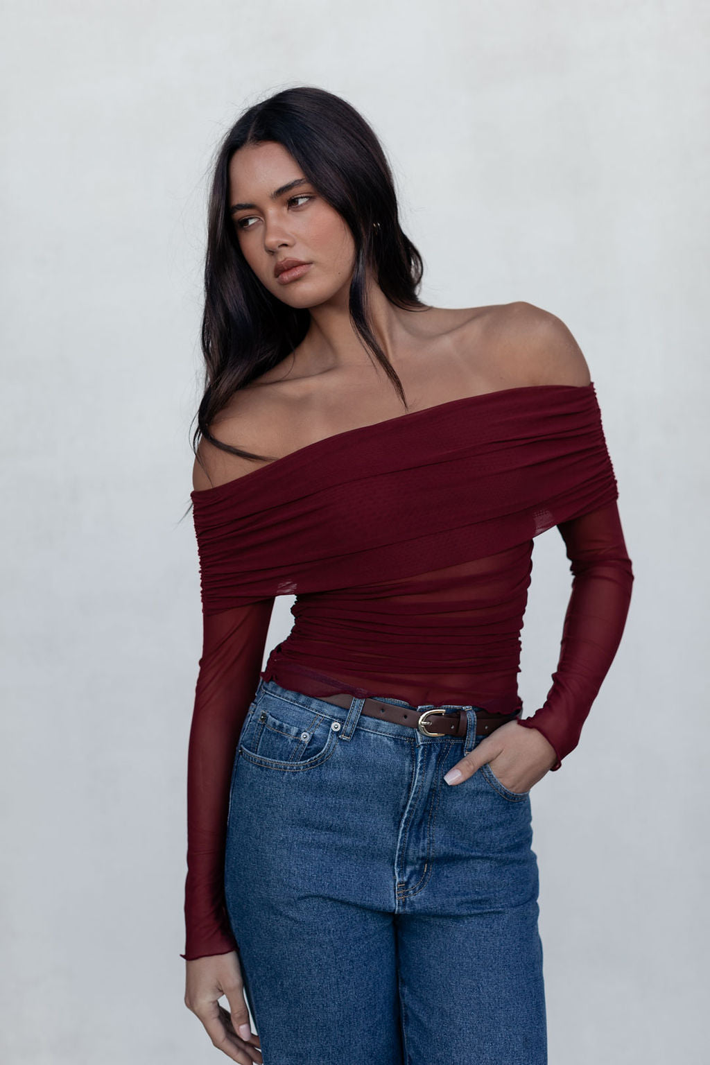 Woman wearing a burgundy off-shoulder top and blue jeans against a plain background