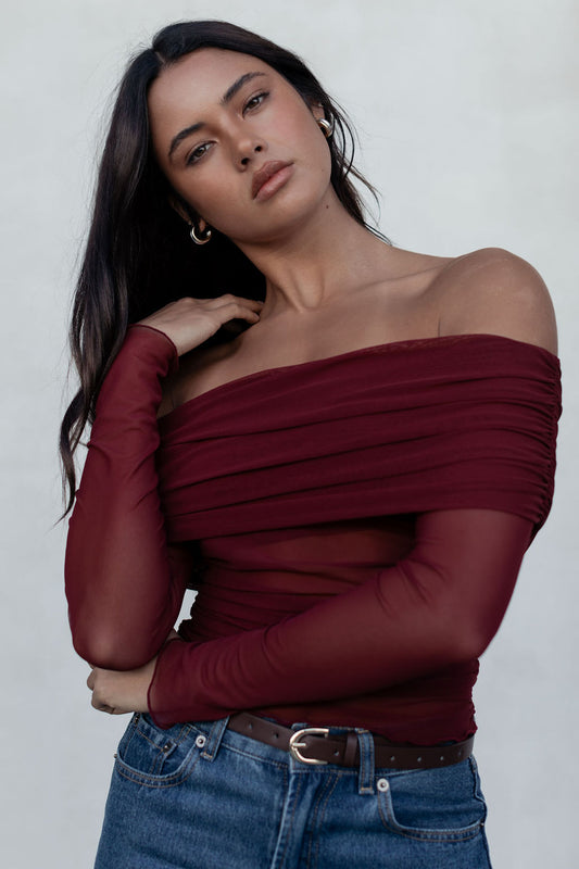 Woman wearing a burgundy off-shoulder top and blue jeans against a plain background