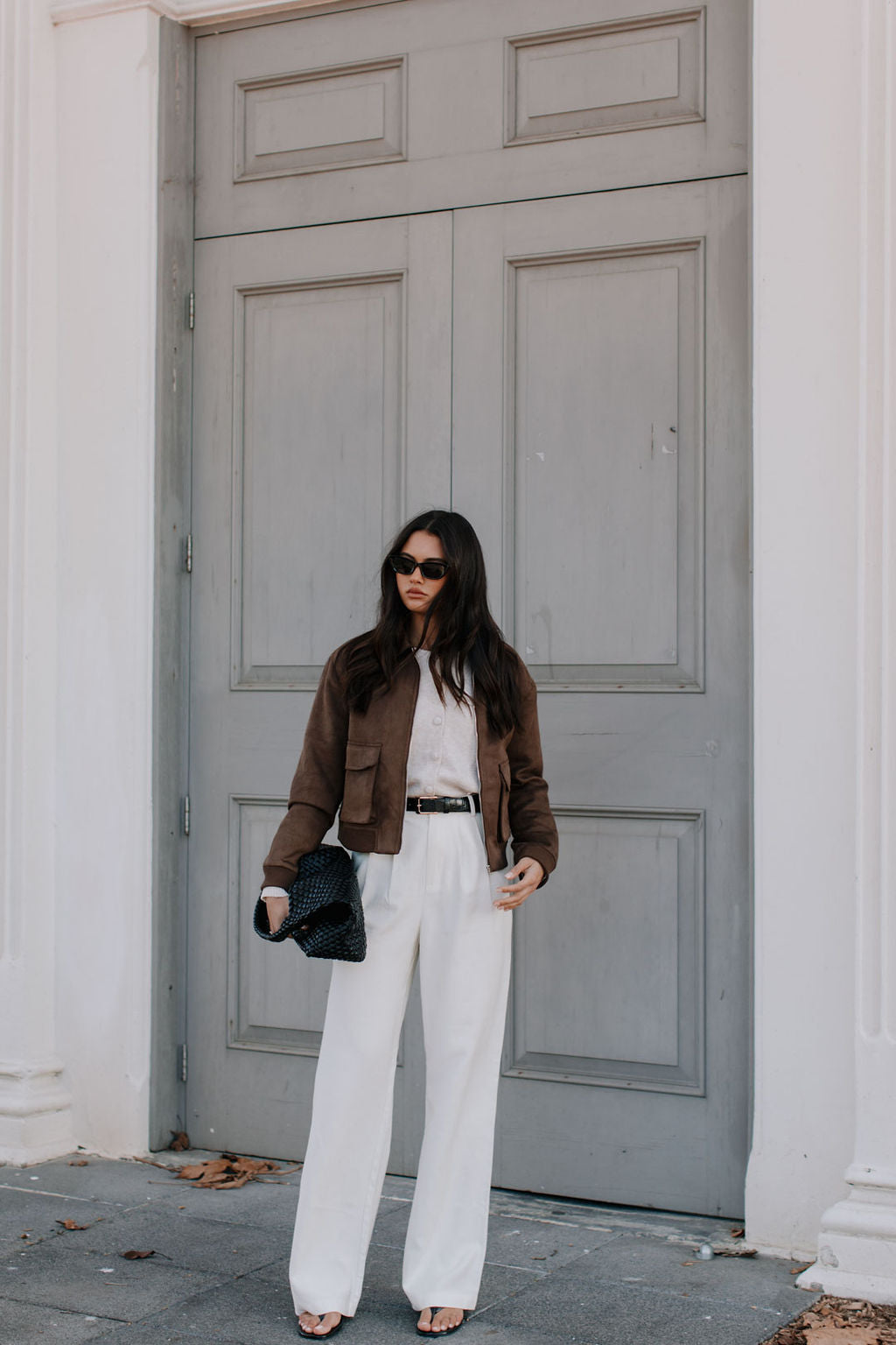 Woman in a brown jacket and white pants standing in front of a gray door.