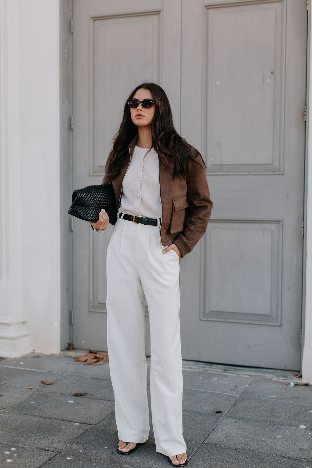 Woman in a stylish outfit with a brown jacket, white shirt, and white pants standing against a gray door.