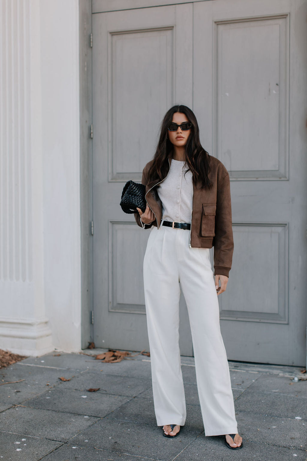 Woman in a stylish outfit with a brown jacket, white pants, and black clutch standing in front of a gray door.
