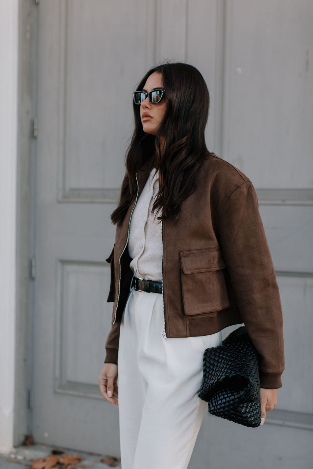 Woman wearing a brown jacket and white pants against a gray wall.