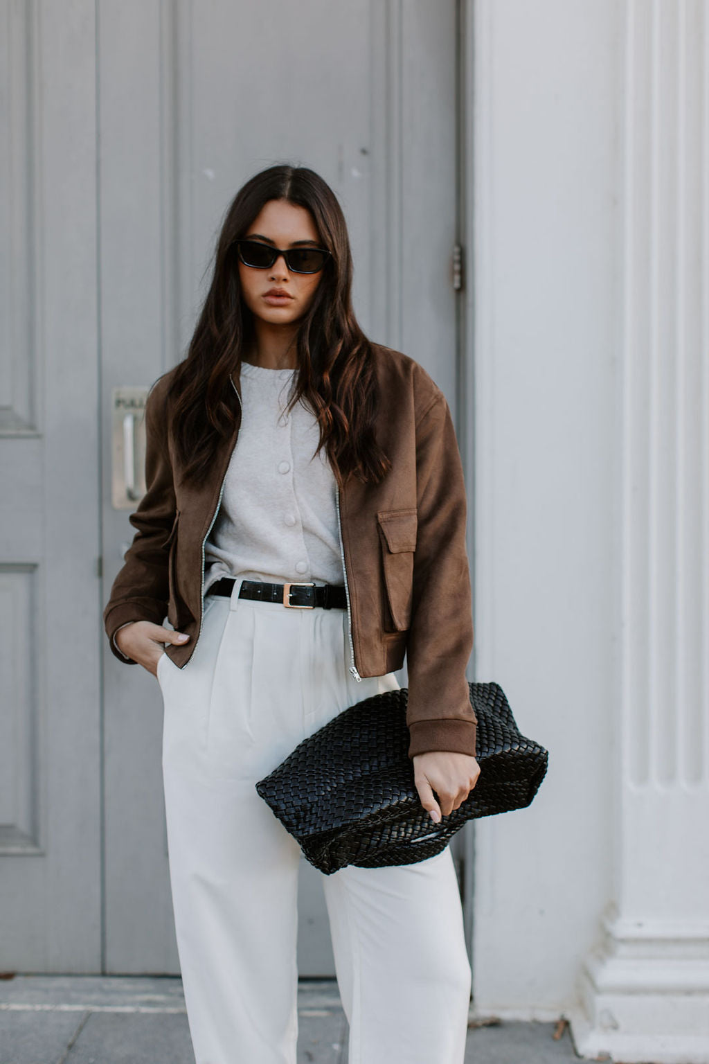 Woman in brown jacket and white pants holding a black bag against a gray door.