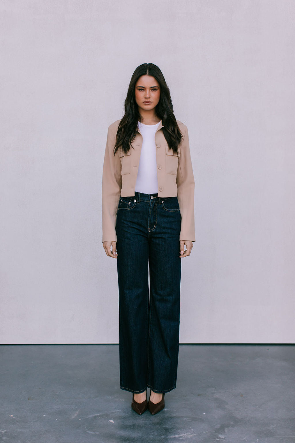 Woman wearing a beige jacket, white shirt, and dark jeans against a plain background