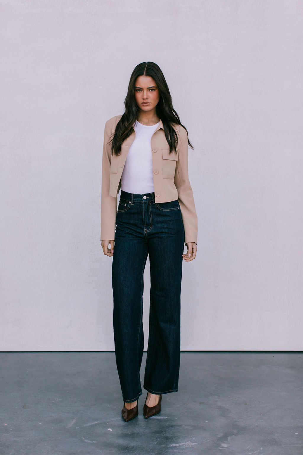 Woman wearing a beige jacket, white top, and dark jeans against a plain background