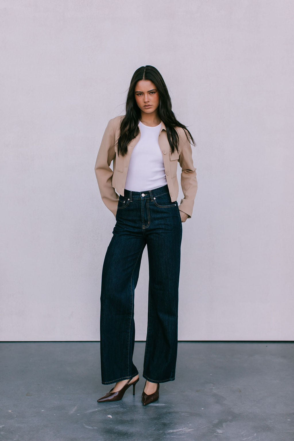 Woman wearing a beige jacket, white shirt, and dark jeans against a plain background