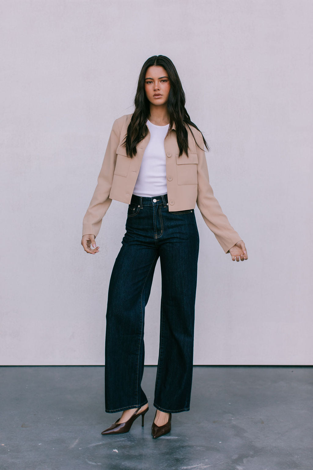 Woman wearing a beige jacket, white top, and dark jeans against a plain background