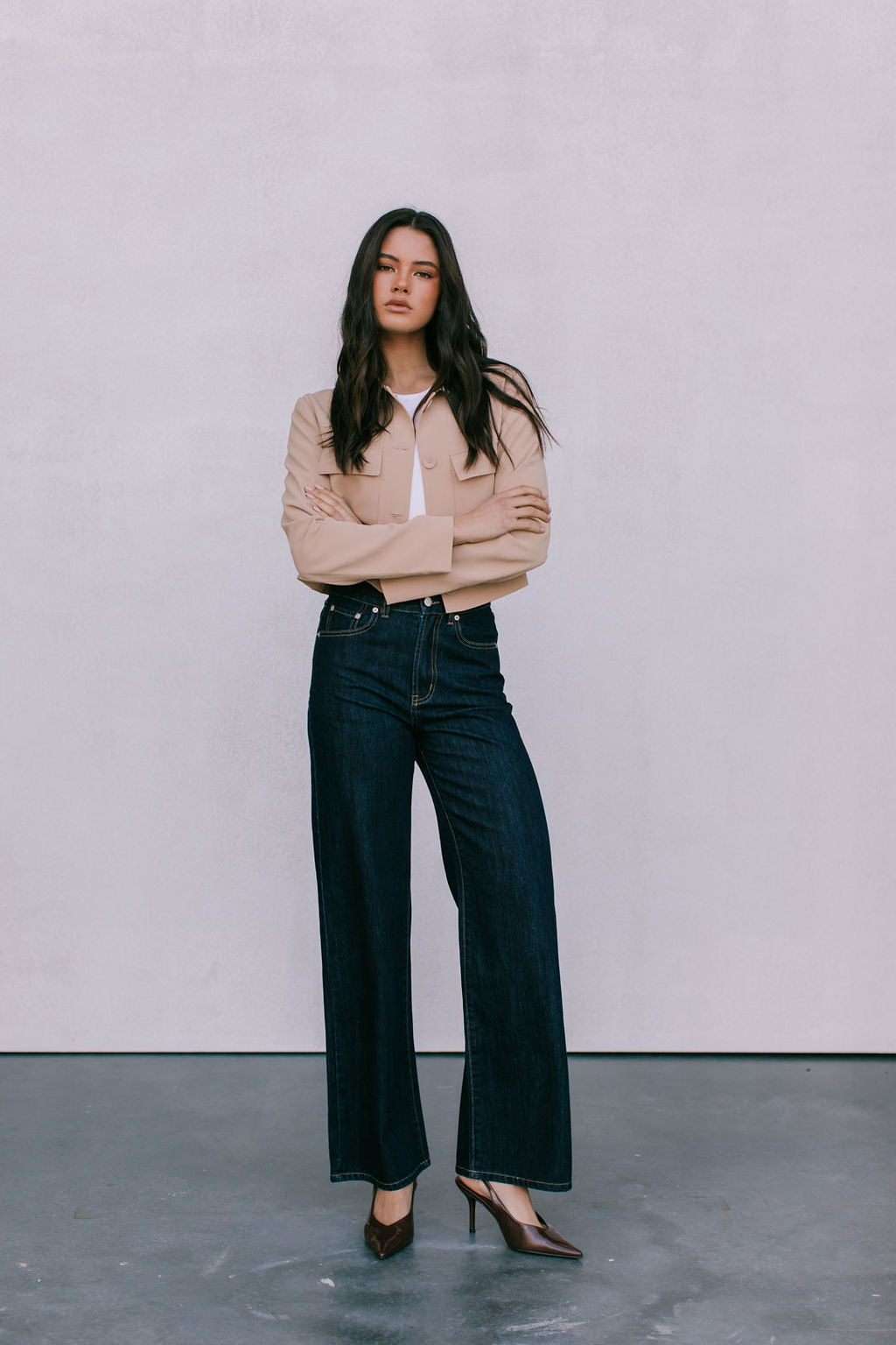 Woman wearing a beige jacket and dark jeans standing against a plain background