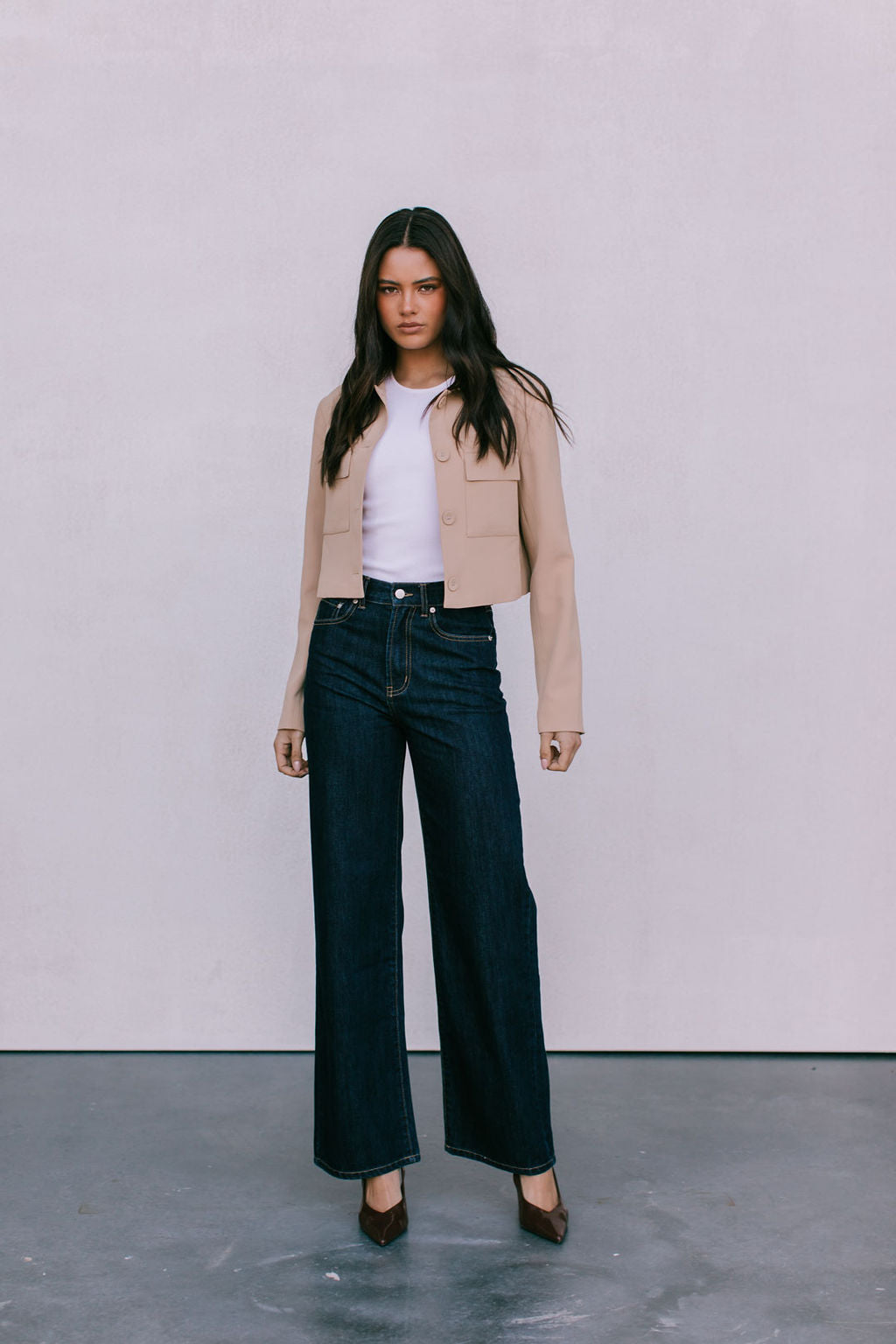 Woman wearing a beige jacket, white shirt, and dark jeans against a plain background