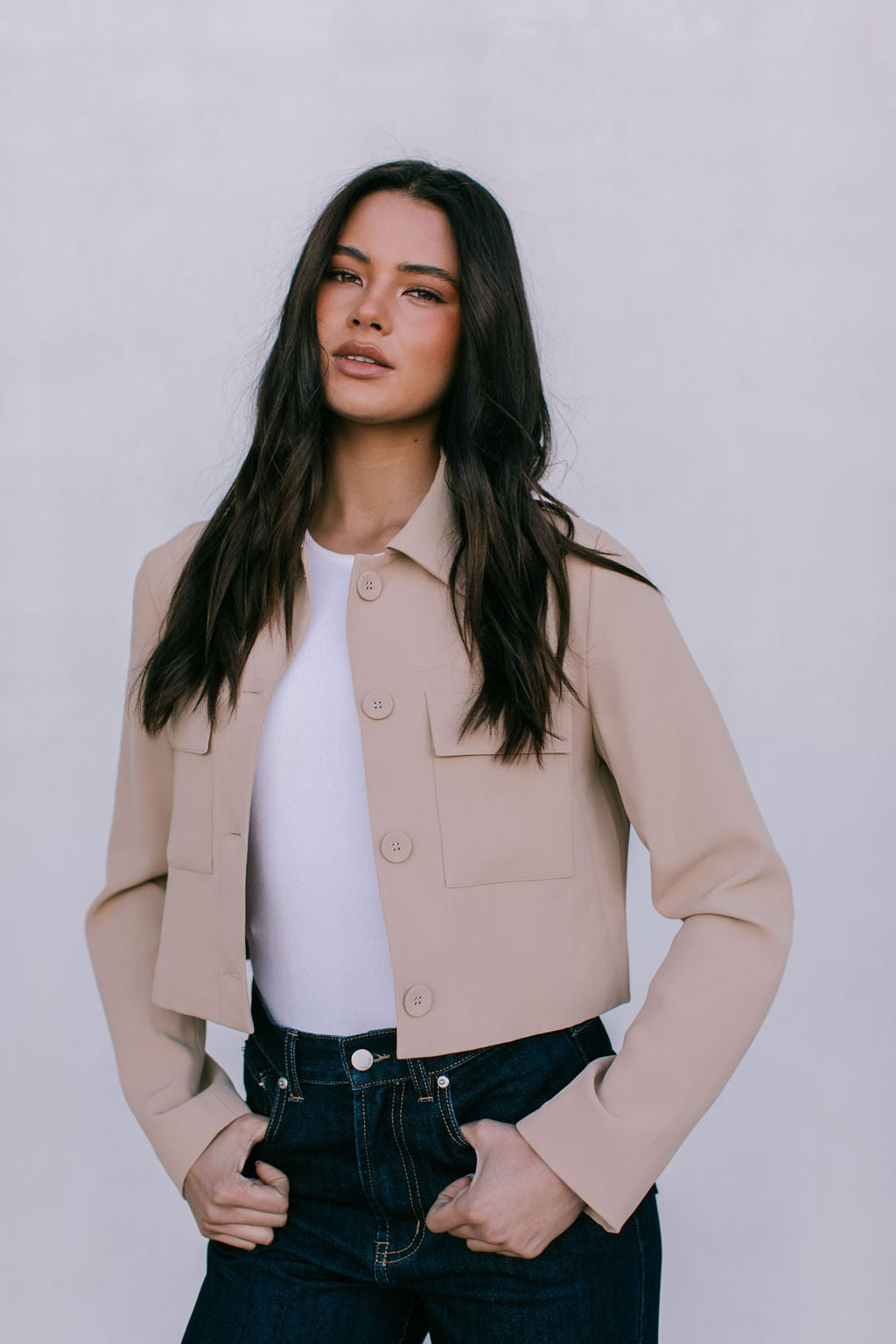 Woman wearing a beige jacket over a white shirt and dark jeans against a plain background