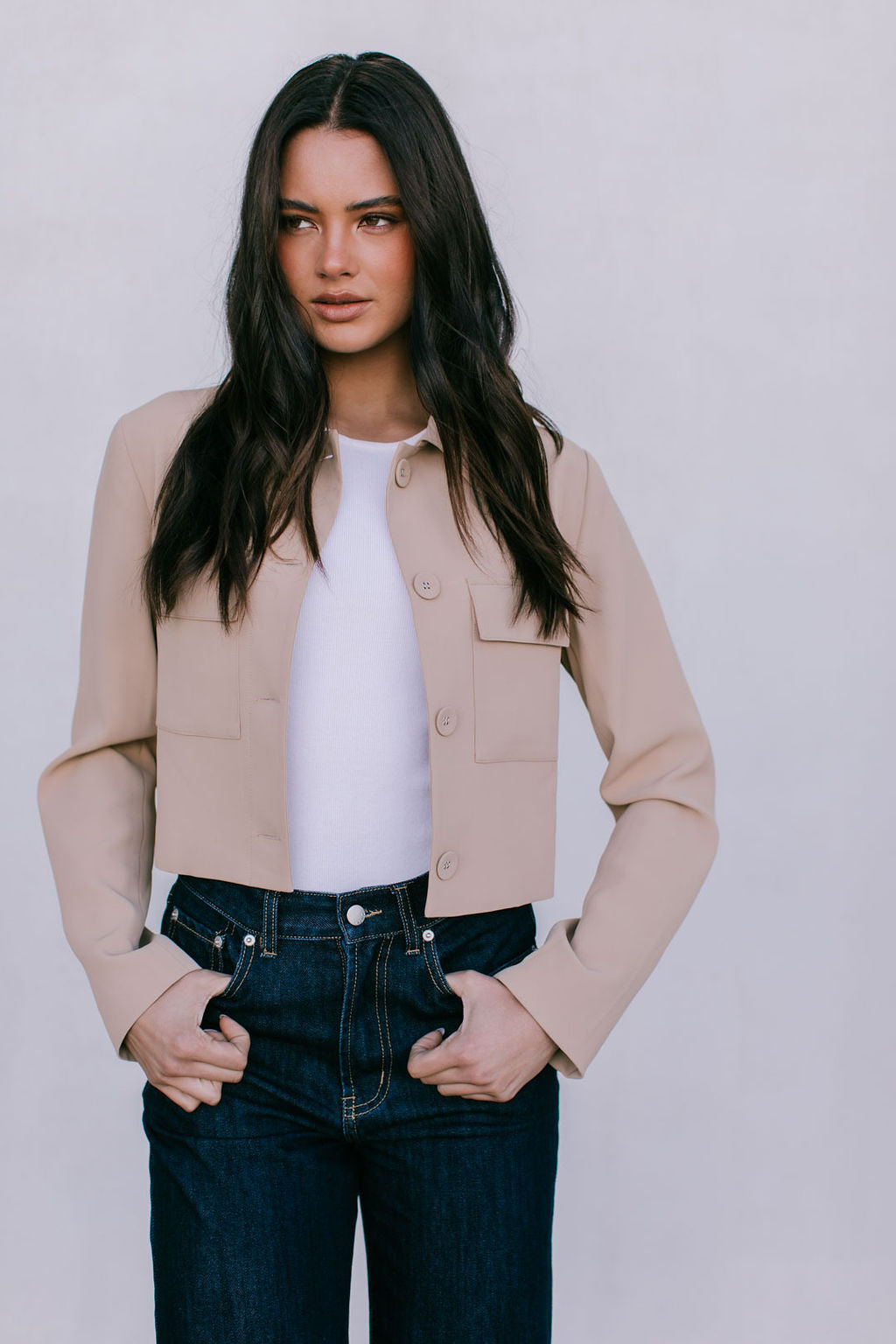 Woman wearing a beige jacket over a white shirt and dark jeans against a plain background