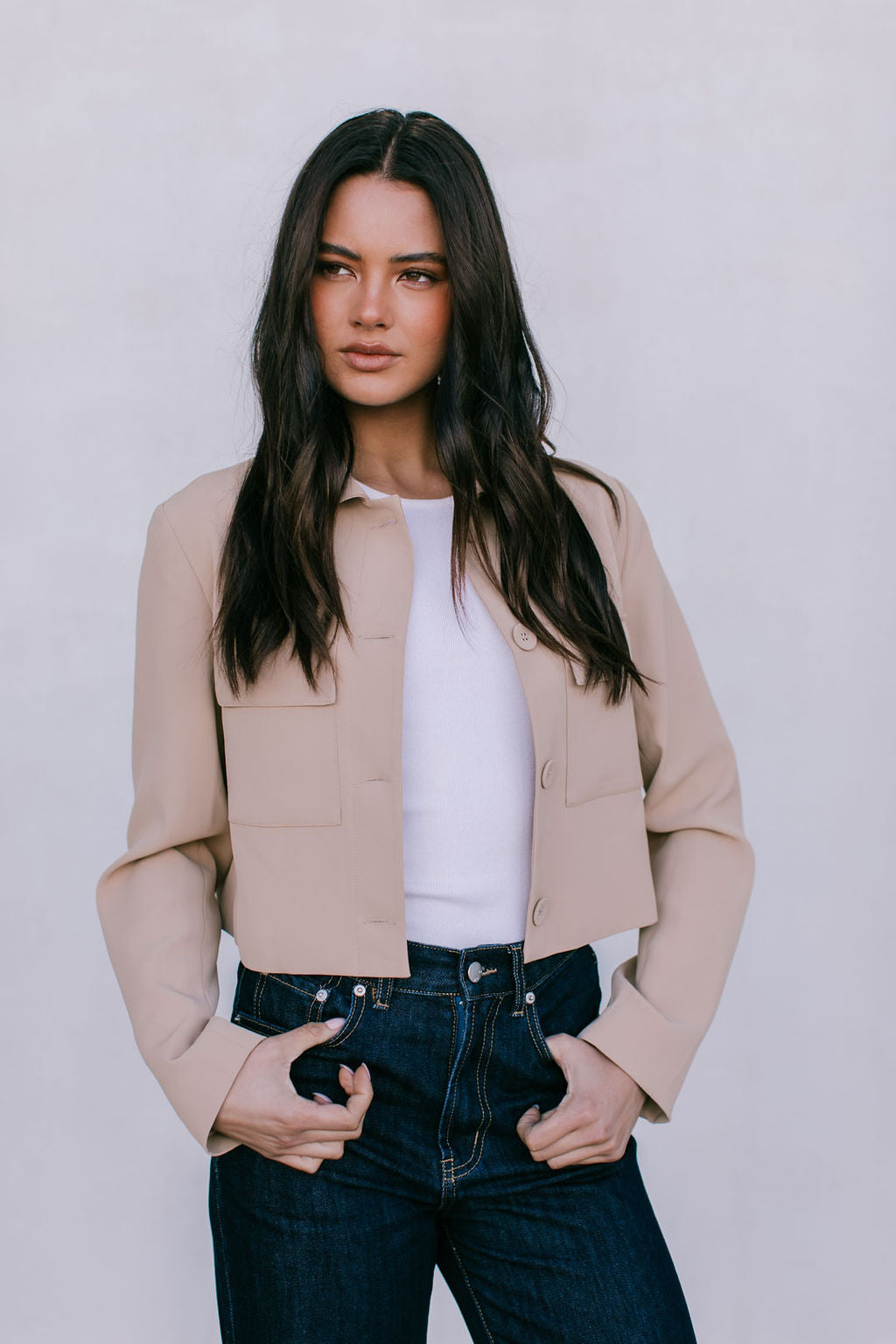 Woman wearing a beige jacket over a white shirt and dark jeans on a plain background
