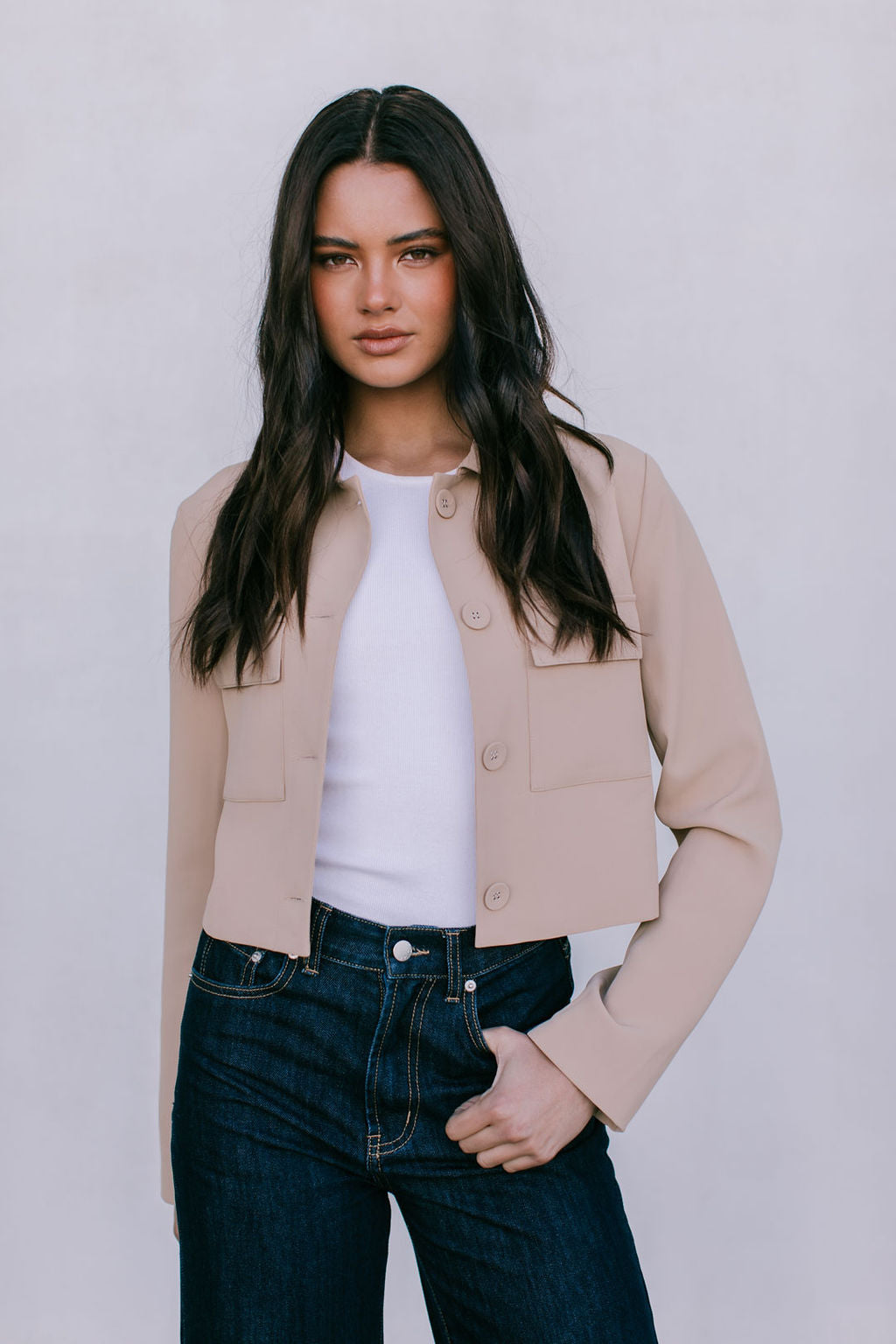 Woman wearing a beige jacket over a white shirt and dark jeans on a plain background