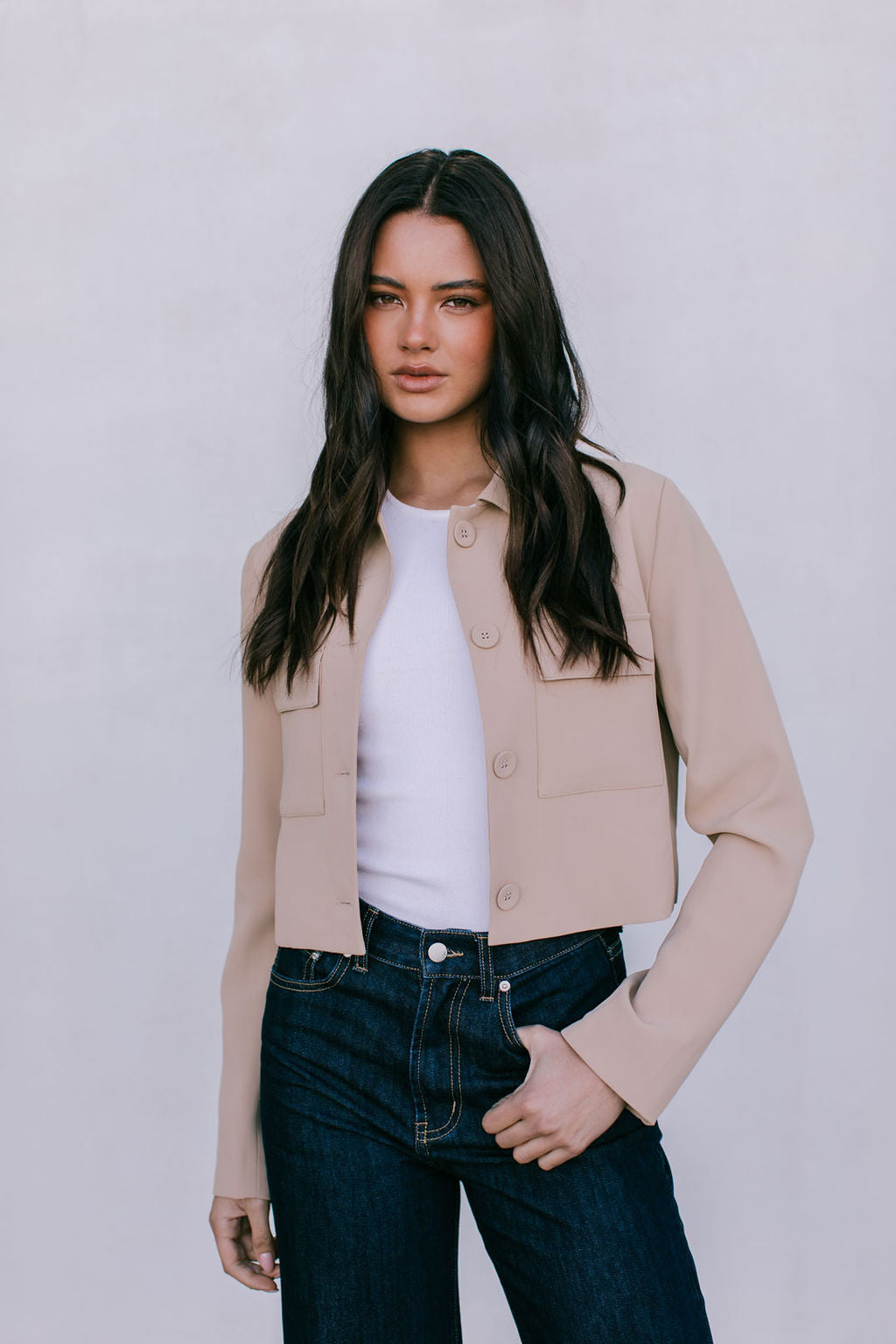Woman wearing a beige jacket, white shirt, and dark jeans against a plain background