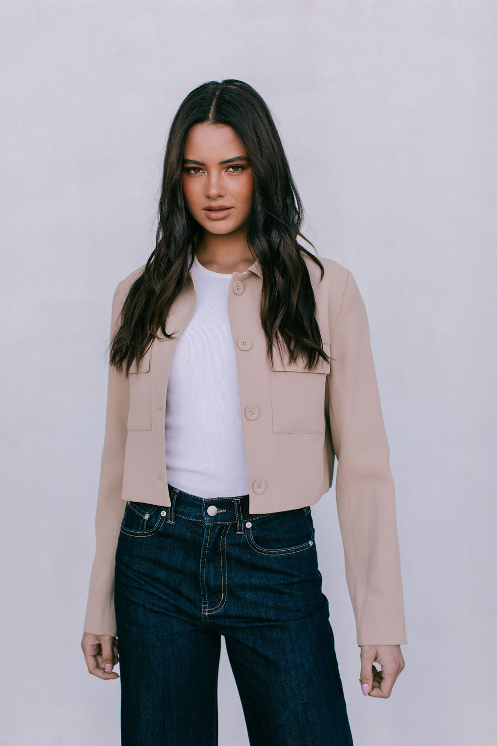 Woman wearing a beige jacket, white shirt, and dark jeans against a plain background