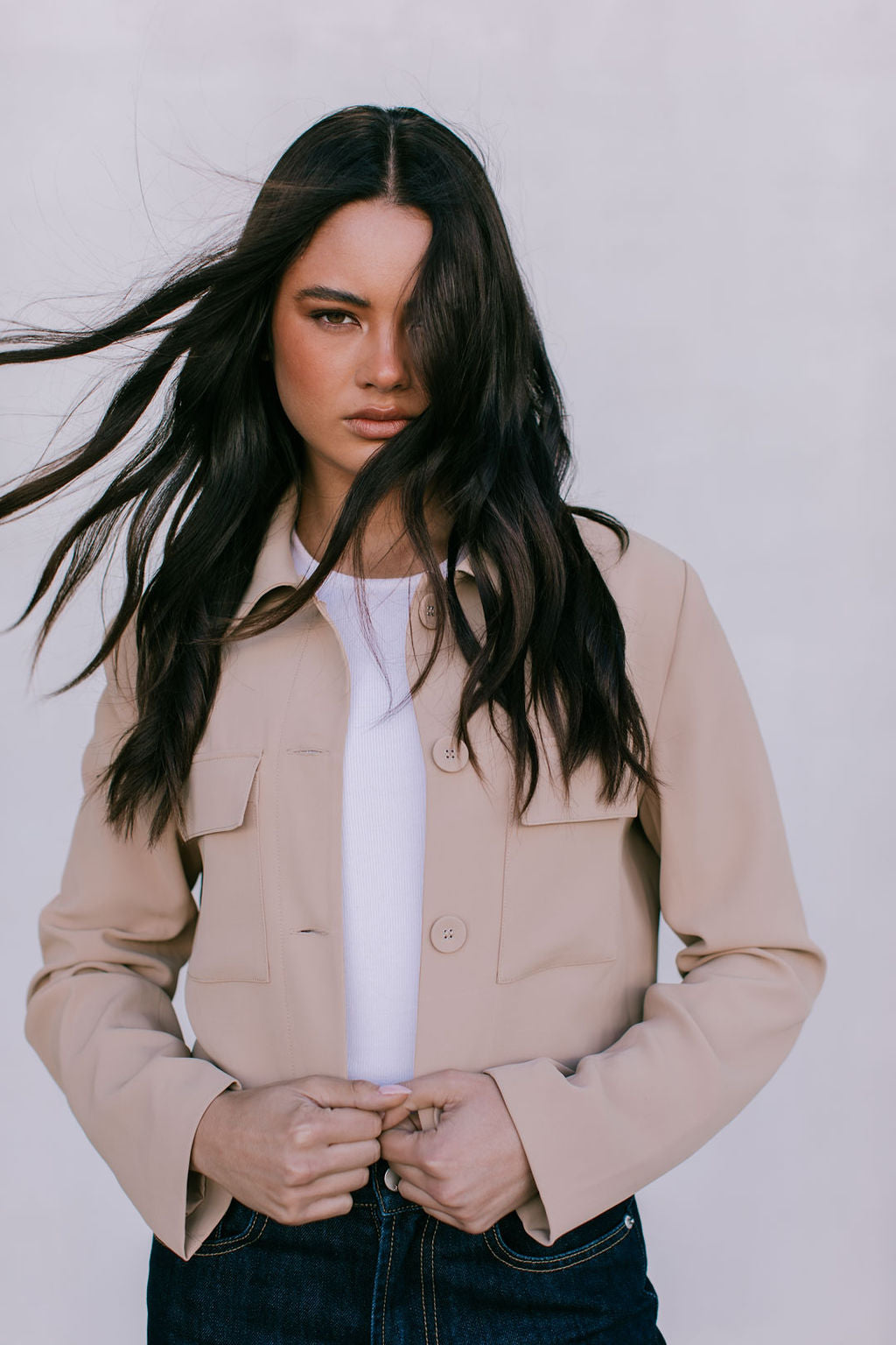 Woman wearing a beige jacket over a white shirt with long dark hair against a plain background