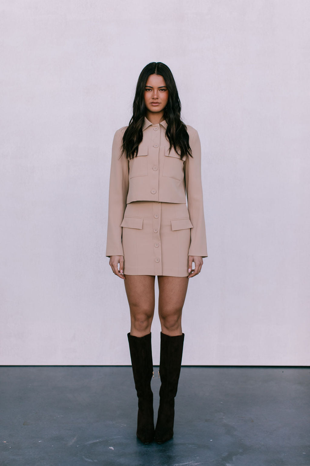 Woman wearing a beige outfit with black knee-high boots on a plain background