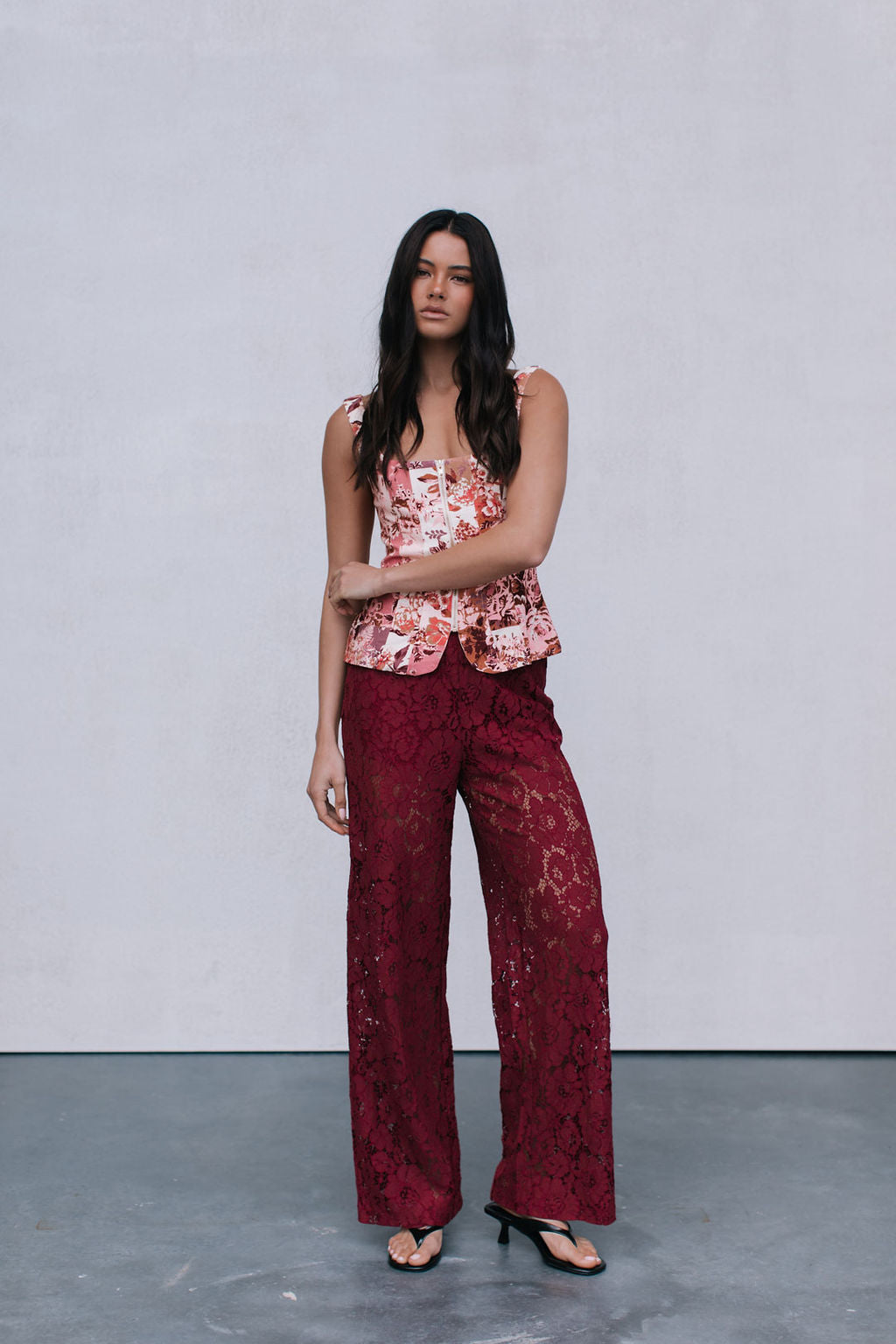 Woman wearing a floral top and burgundy lace pants against a plain background