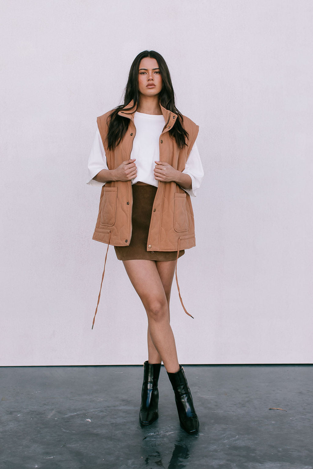 Woman wearing a beige vest over a white shirt and brown skirt with black boots on a plain background