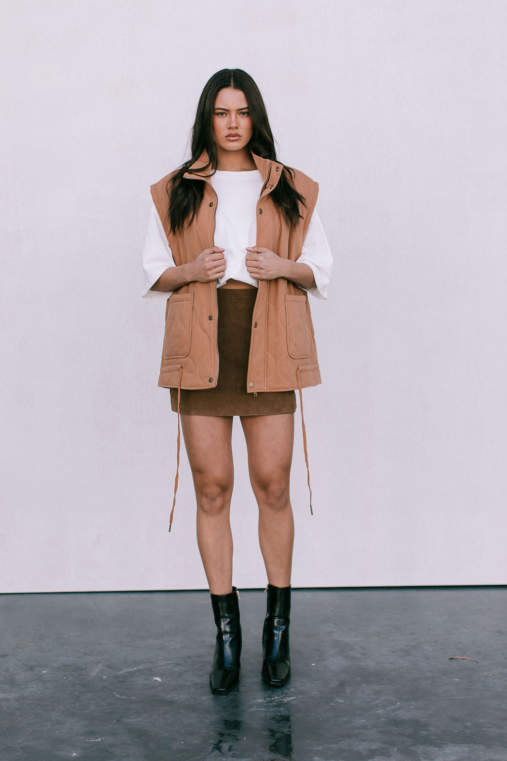 Woman wearing a tan vest over a white shirt and brown skirt with black boots on a plain background