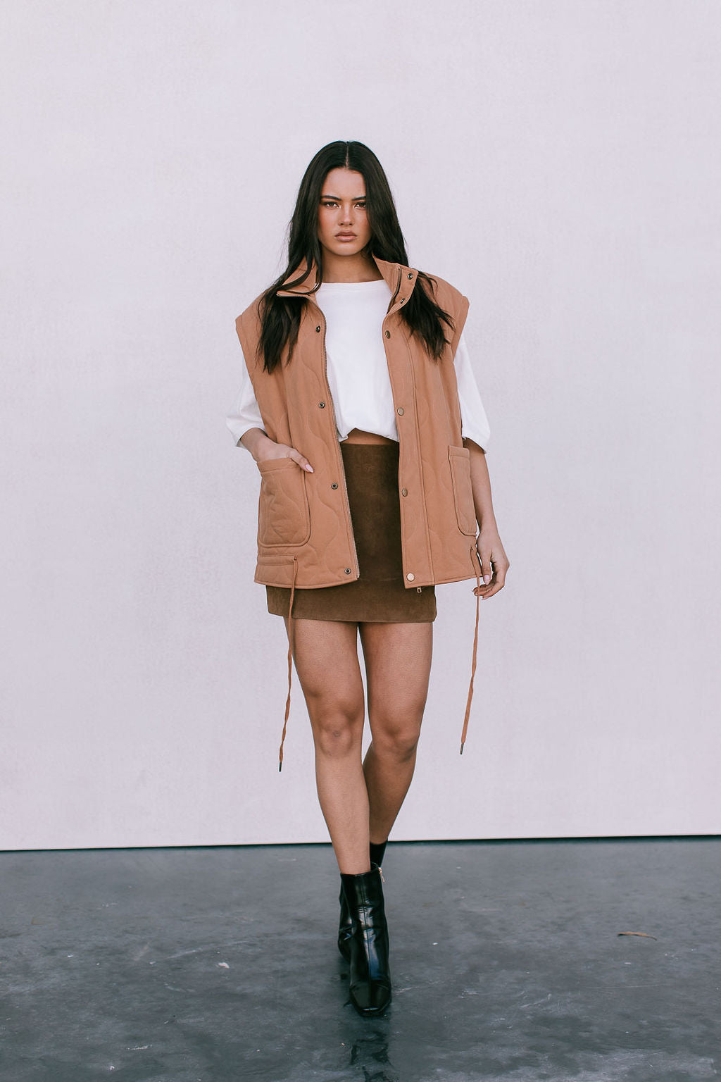 Woman wearing a tan quilted vest, white top, brown skirt, and black boots on a plain background