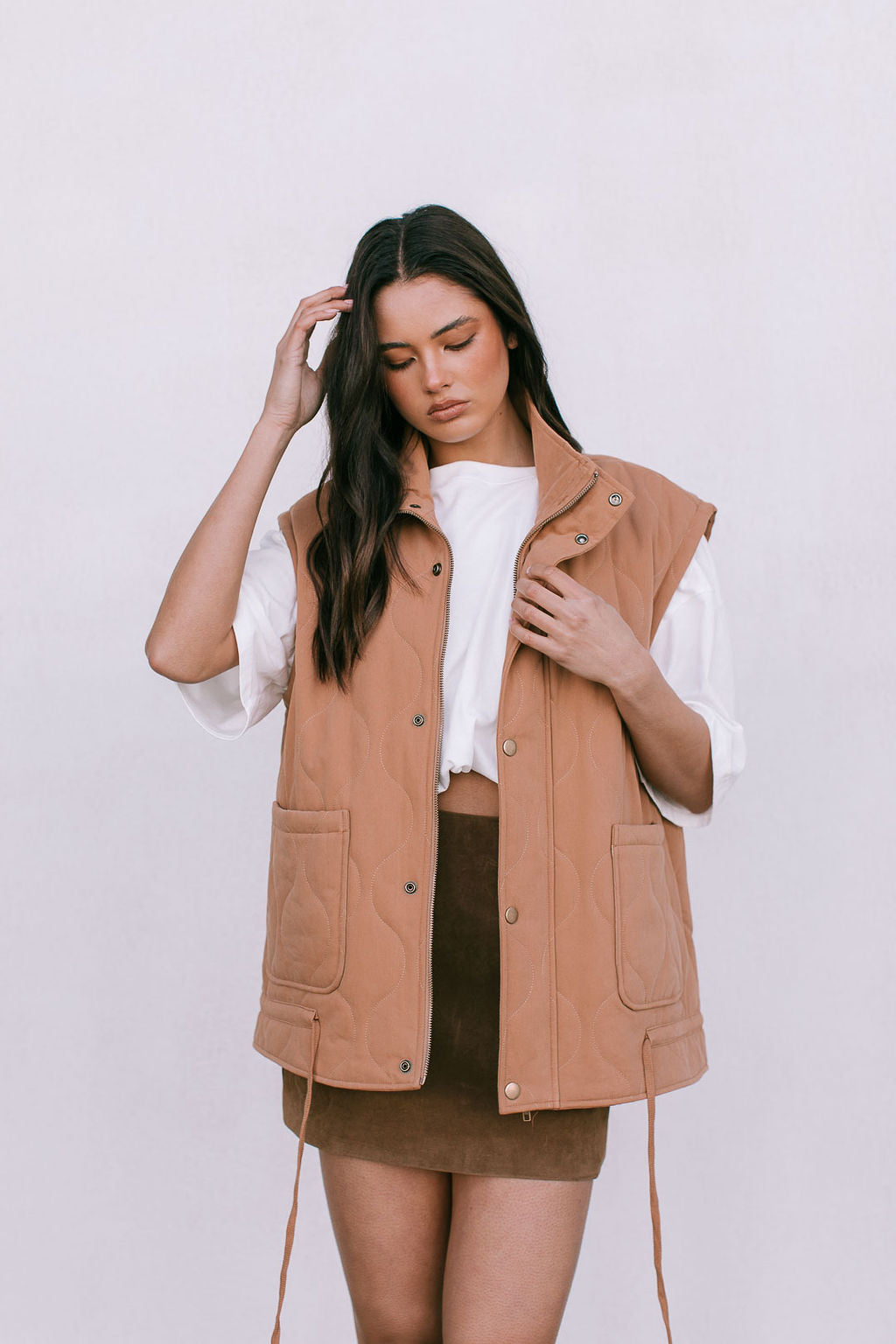 Woman wearing a tan vest over a white shirt and brown skirt on a white background