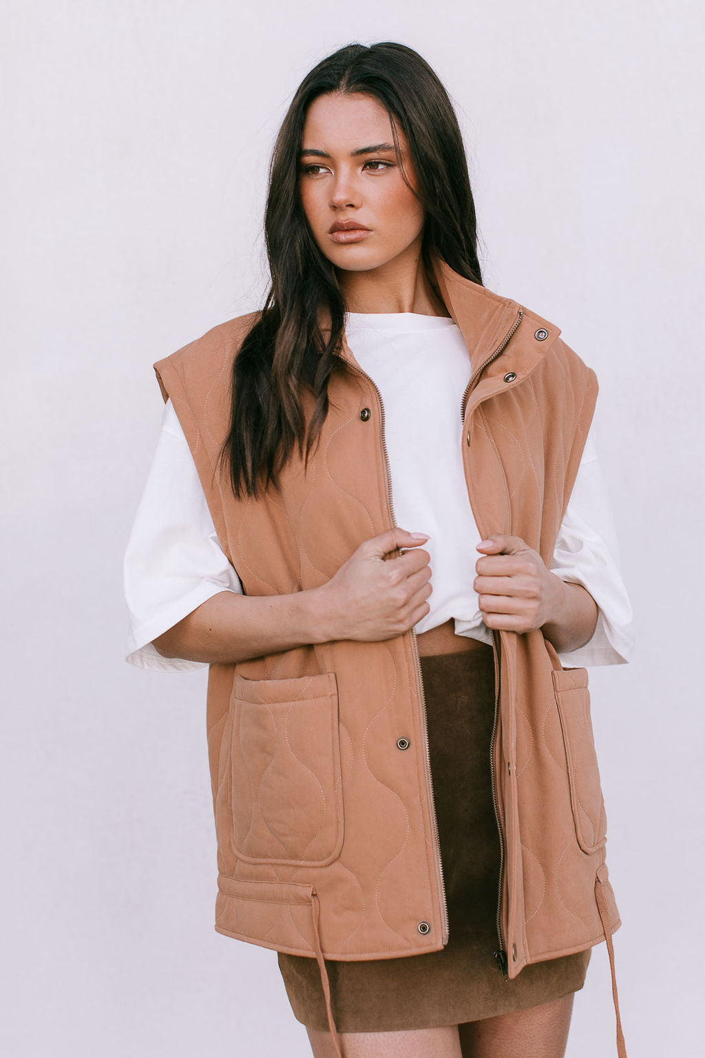 Woman wearing a tan vest over a white shirt and brown skirt on a plain background