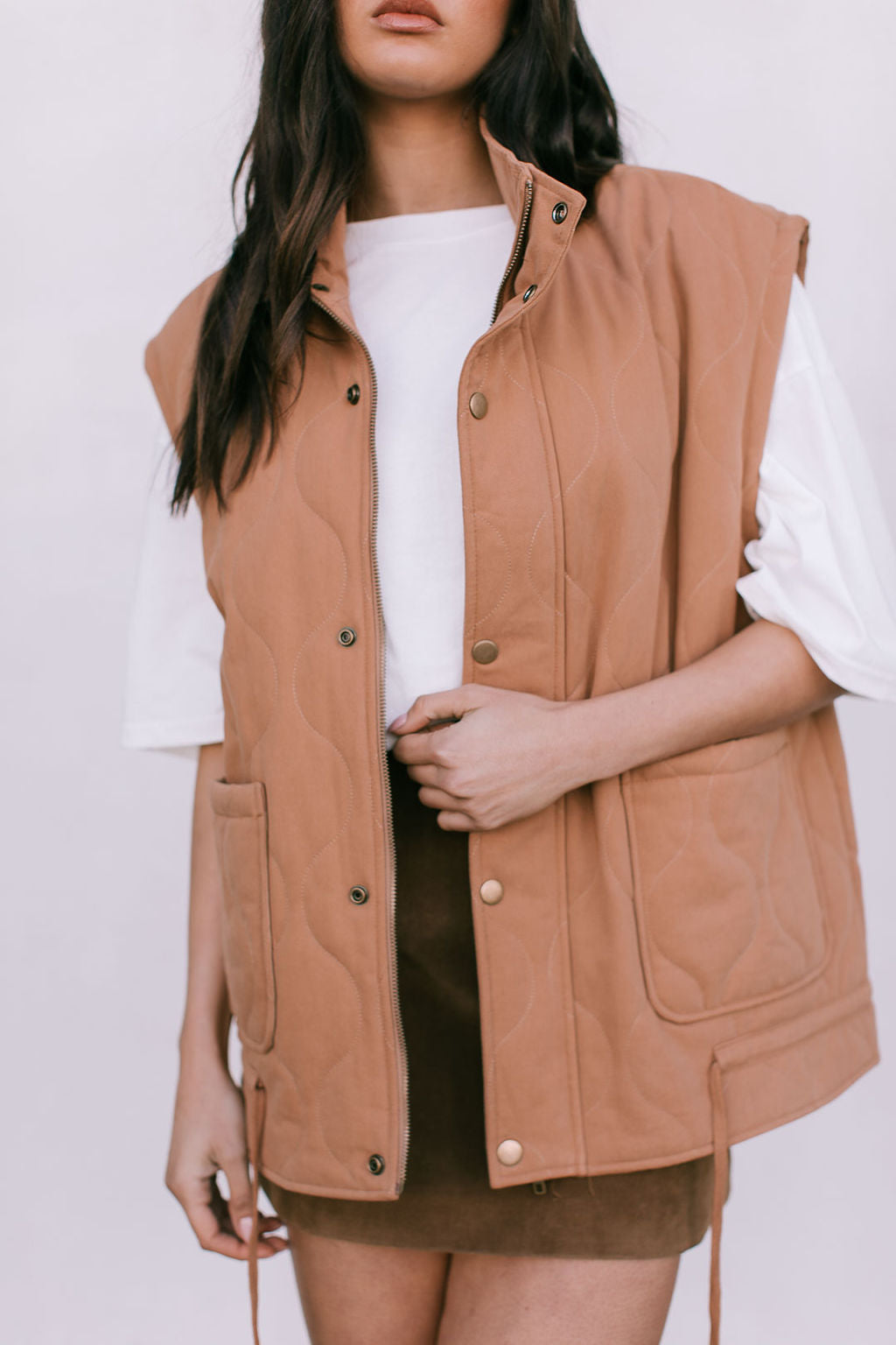 Person wearing a brown vest over a white shirt and black skirt on a plain background
