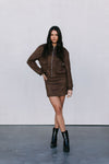 Woman wearing a brown jacket and skirt set with black boots on a plain background