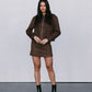 Woman wearing a brown outfit with a jacket and skirt, standing against a plain background.