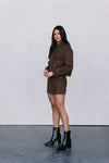 Woman wearing a brown jacket and skirt with black boots against a white wall.