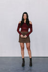 Woman wearing a burgundy off-shoulder top and brown skirt with black boots against a plain background